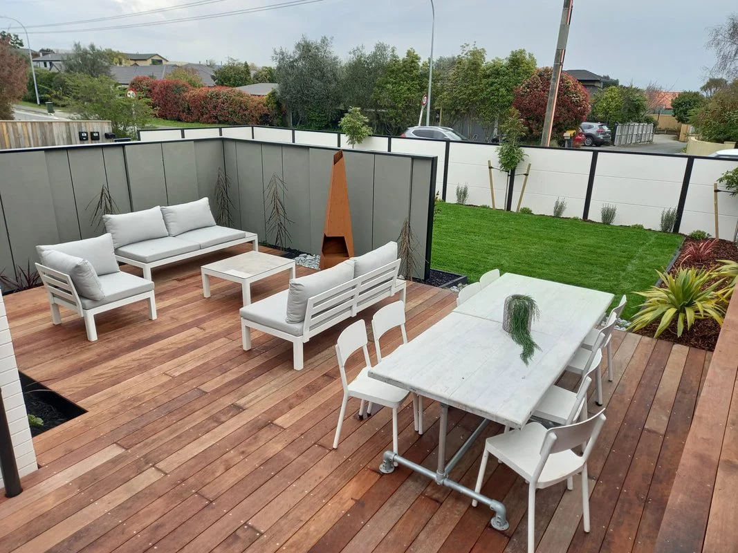 A modern outdoor patio with wooden flooring, white outdoor furniture, including chairs, sofas, and a marble dining table with chairs, surrounded by a garden with green grass and plants, enclosed by a grey and white privacy fence, with colored bushes 