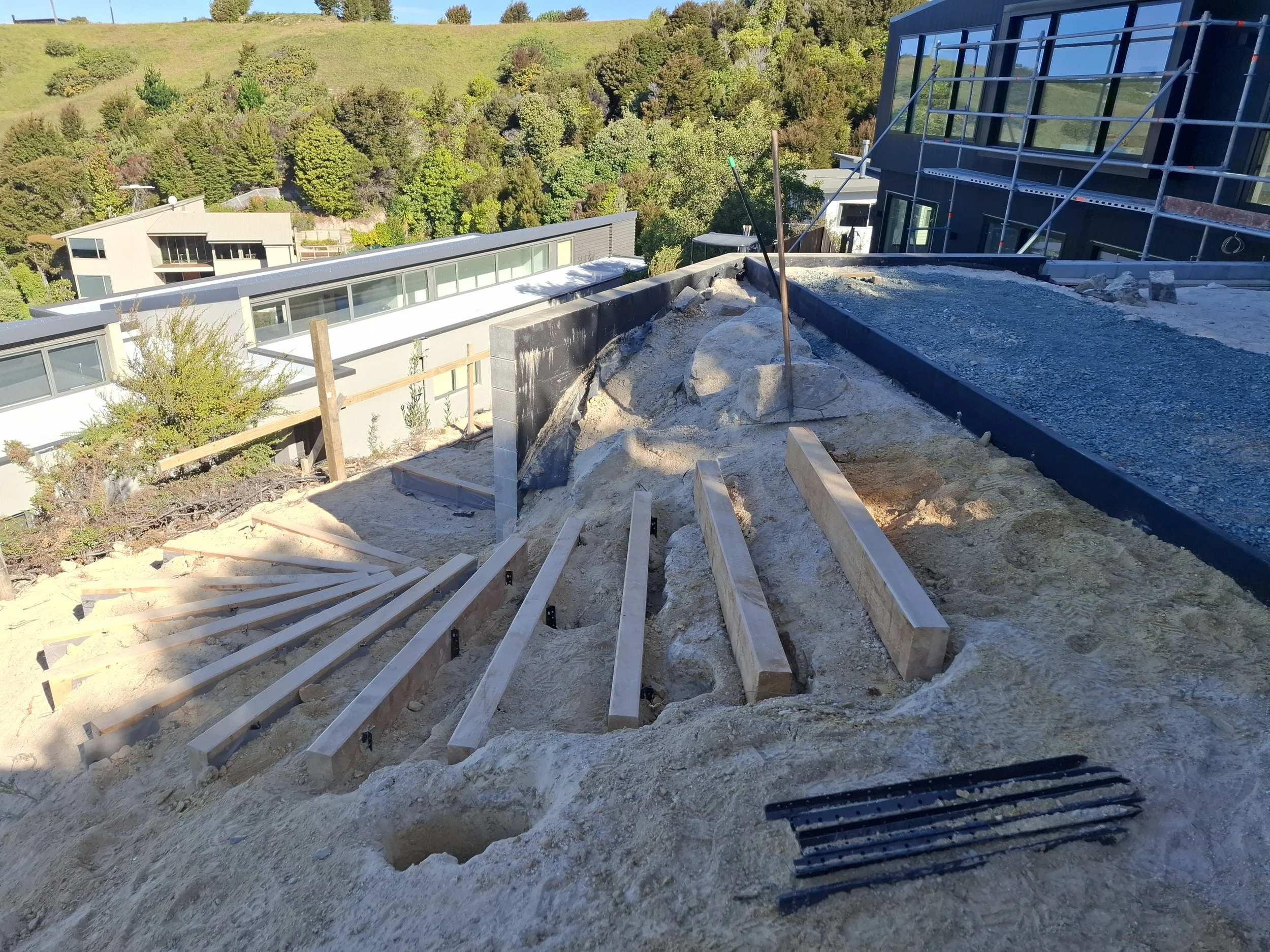 Construction site on a hillside with a building in progress, showing wooden framing and gravel foundation, surrounded by greenery and trees.
