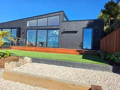 Modern black house with large glass windows and sliding glass doors, set on a grassy yard with planters and stone pathway, under a bright blue sky.