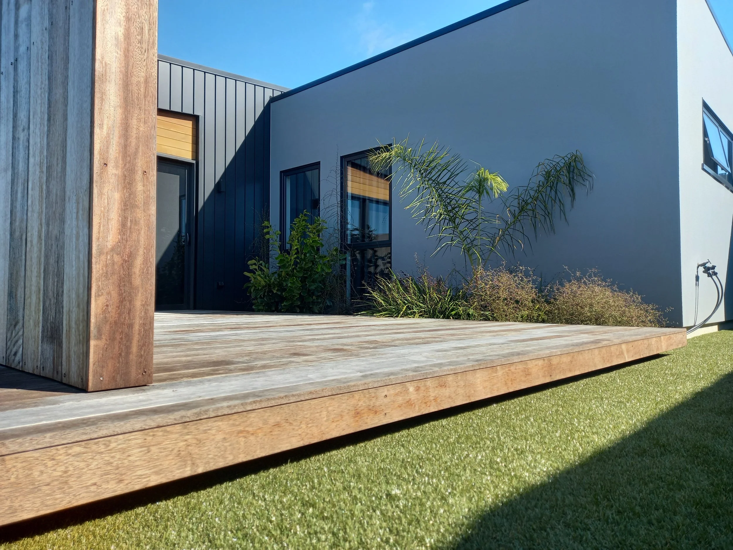 A modern house with a flat roof, gray and white exterior walls, large windows with wooden blinds, and a wooden deck. There are plants and a garden with grass in front of the house. The sky is clear and blue.