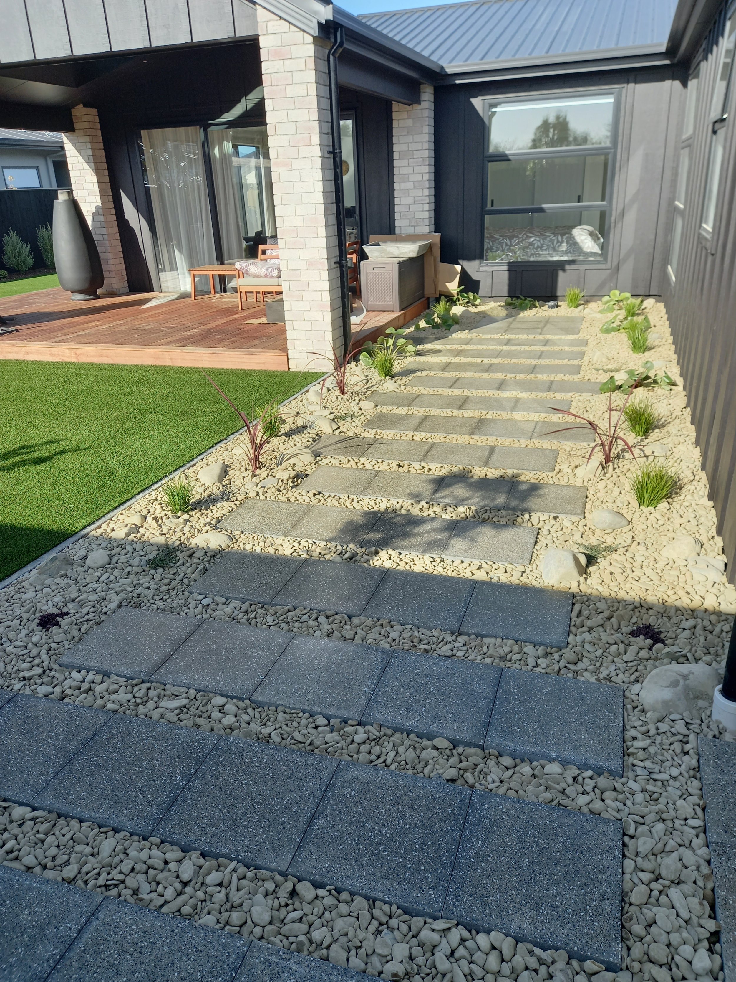 A backyard patio with a wooden deck area, outdoor furniture, and a small pathway made of large square pavers bordered by gravel and sparse plants.