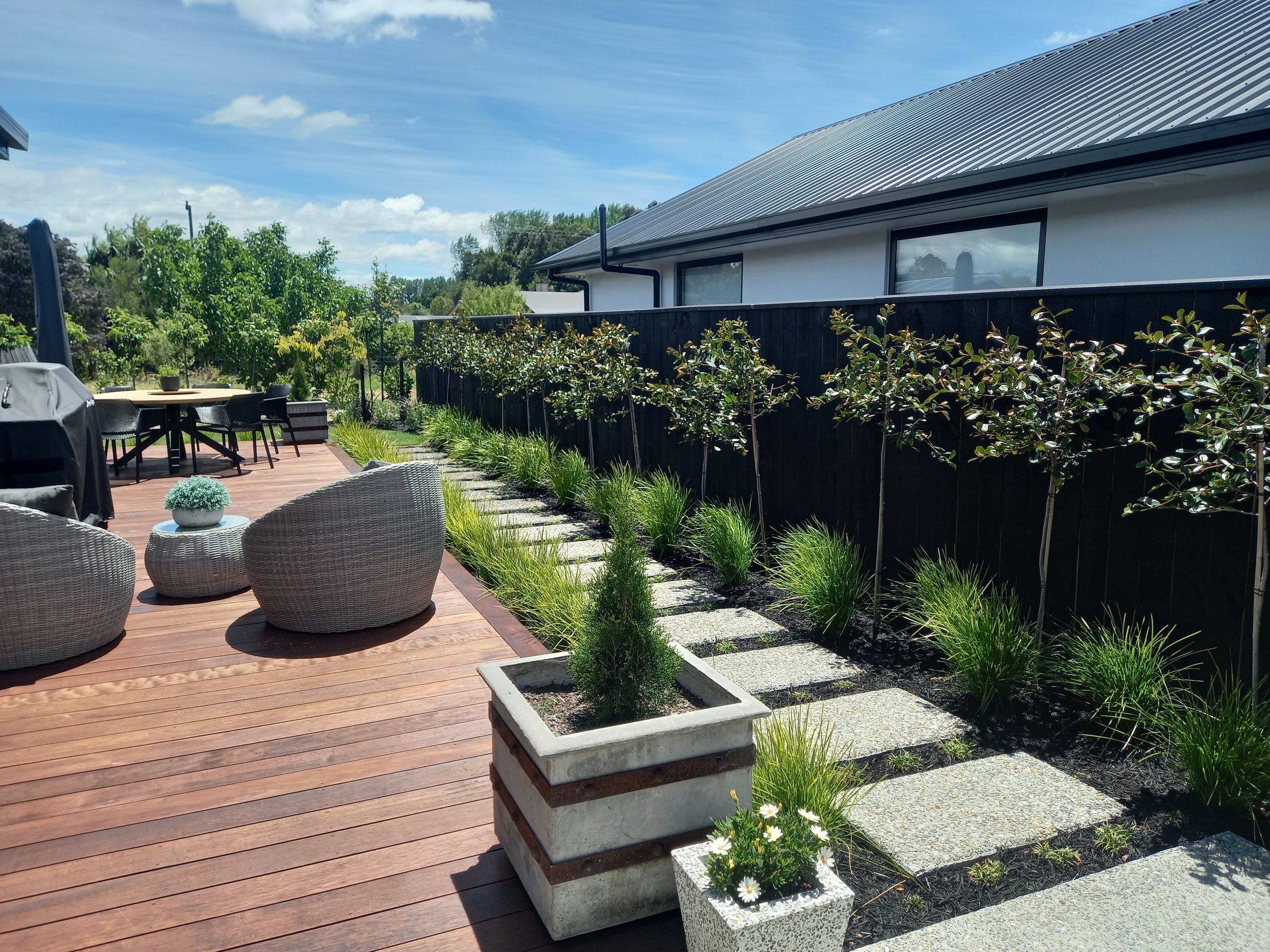 A backyard deck with outdoor furniture, potted plants, and greenery, bordered by a black wooden fence and a white house with large windows under a partly cloudy sky.