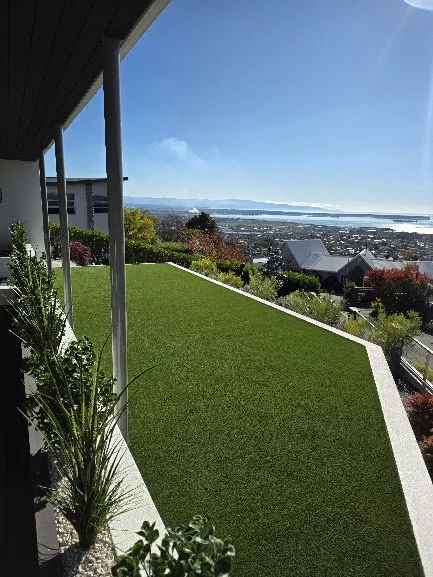 View from a balcony showing a maintained lawn, colorful trees, and a distant cityscape with water under a clear blue sky.