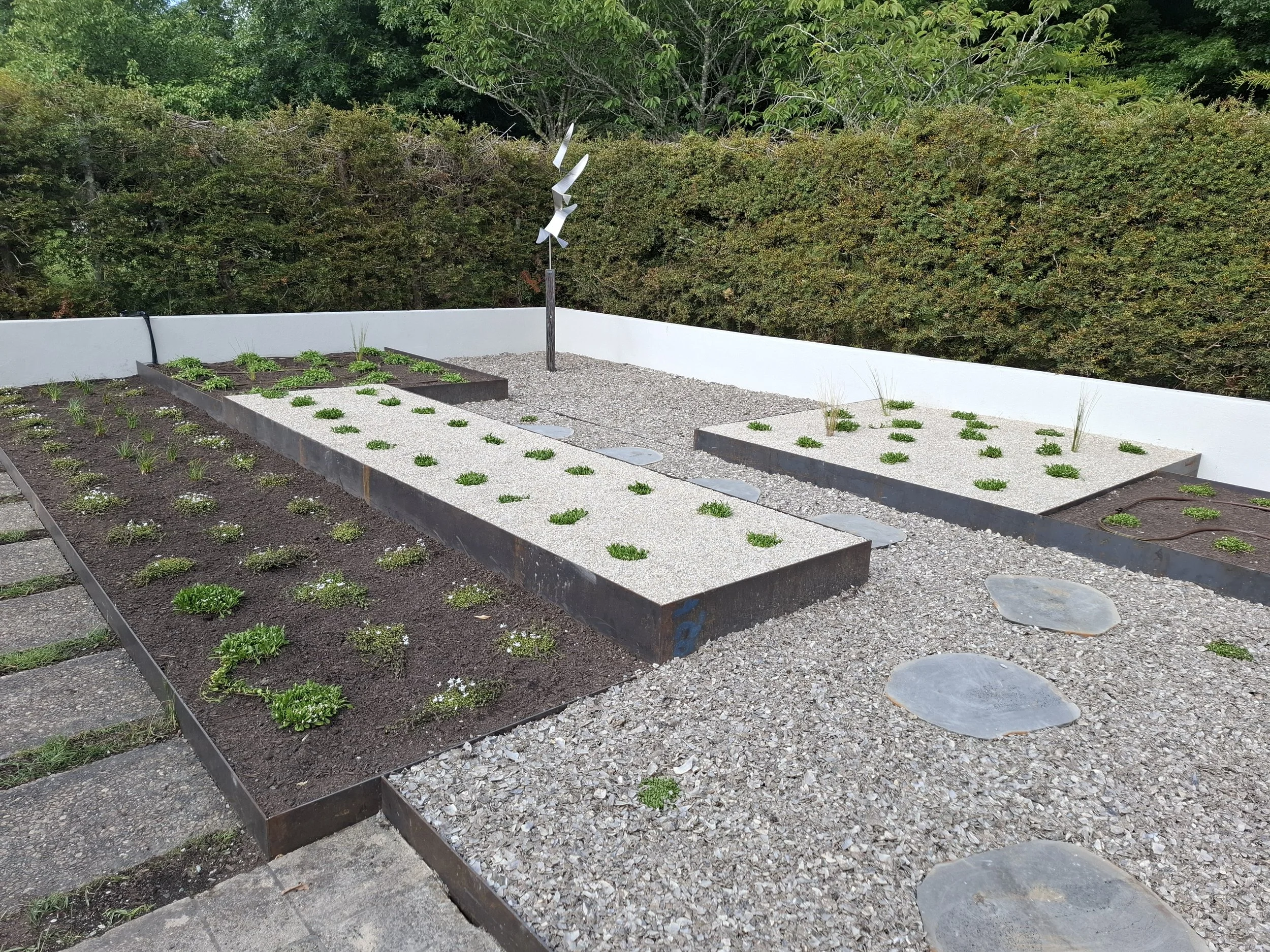 A small garden with young plants growing in various soil beds, surrounded by gravel paths, and a metallic wind sculpture in the background, with dense green bushes and trees behind a white wall.
