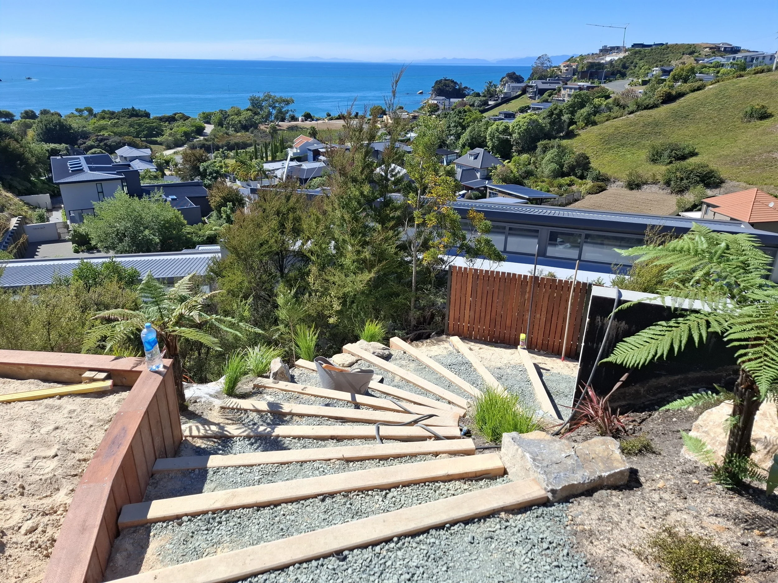 A scenic view of a hillside neighborhood overlooking the ocean with clear blue skies, green trees, and modern houses.