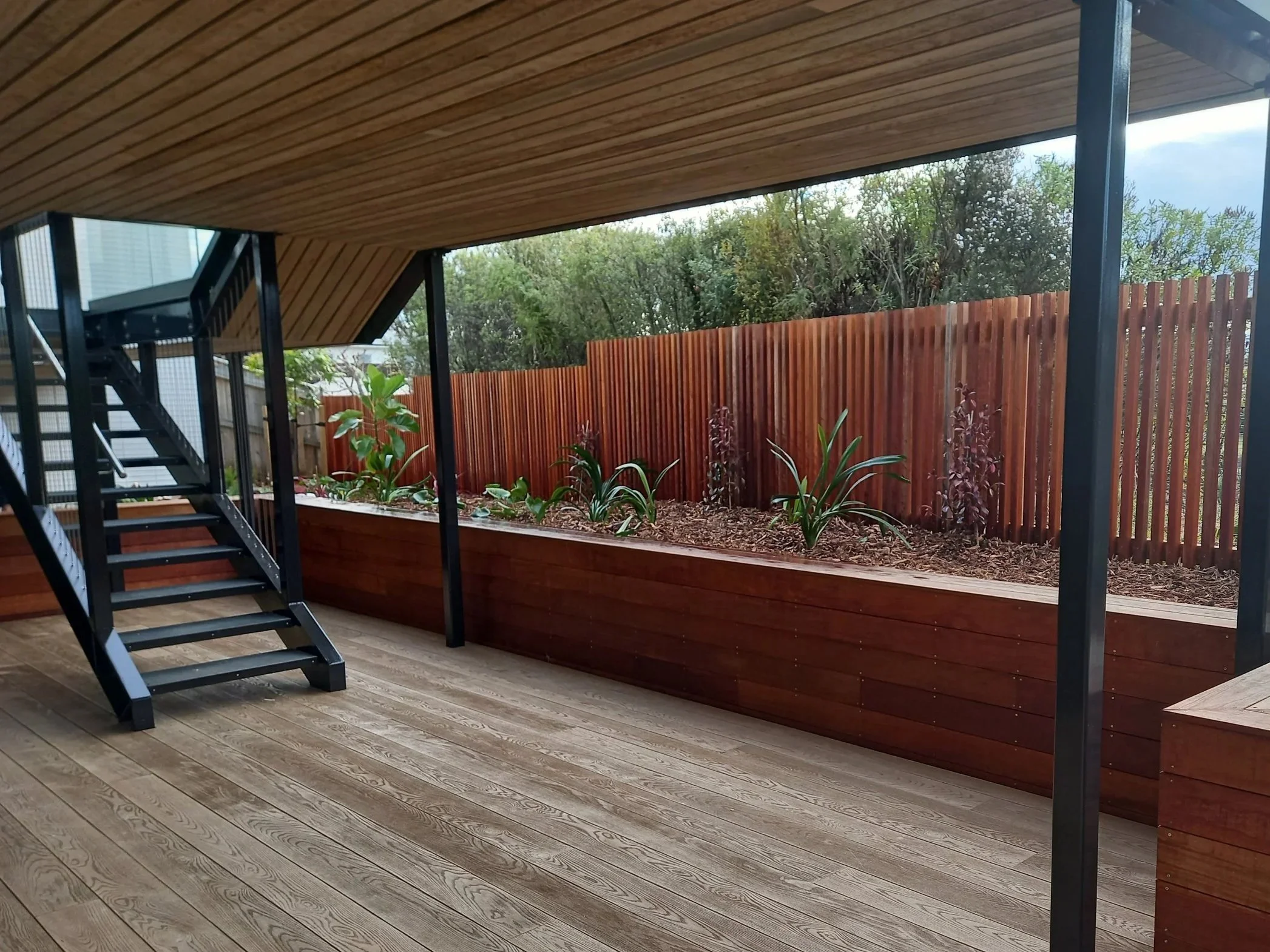 View of a covered outdoor patio area with wooden flooring and ceiling, a black metal staircase, a wood privacy fence, and various plants in a garden bed along the fence, with trees visible in the background.