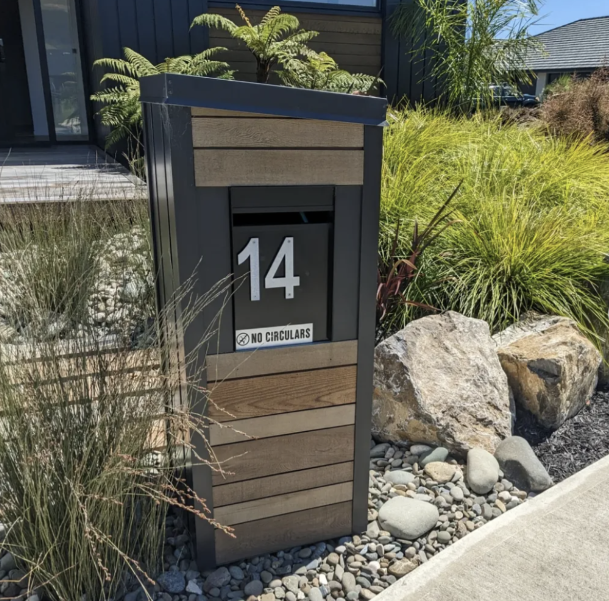 House number 14 mailbox with a black front, wooden panels, and a 'No Circulars' sign, surrounded by landscaped garden with rocks and plants.