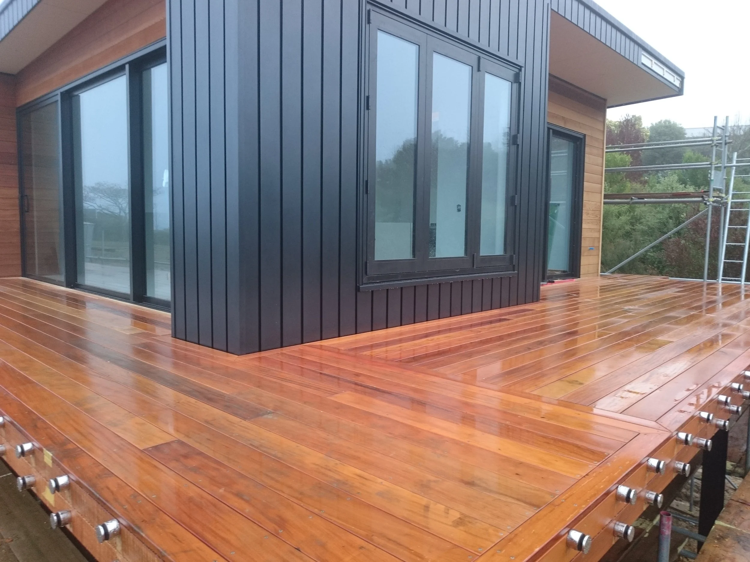 Exterior view of a modern house with large glass sliding doors, black metal siding, and wooden paneling, with scaffolding on the side and a wet wooden deck