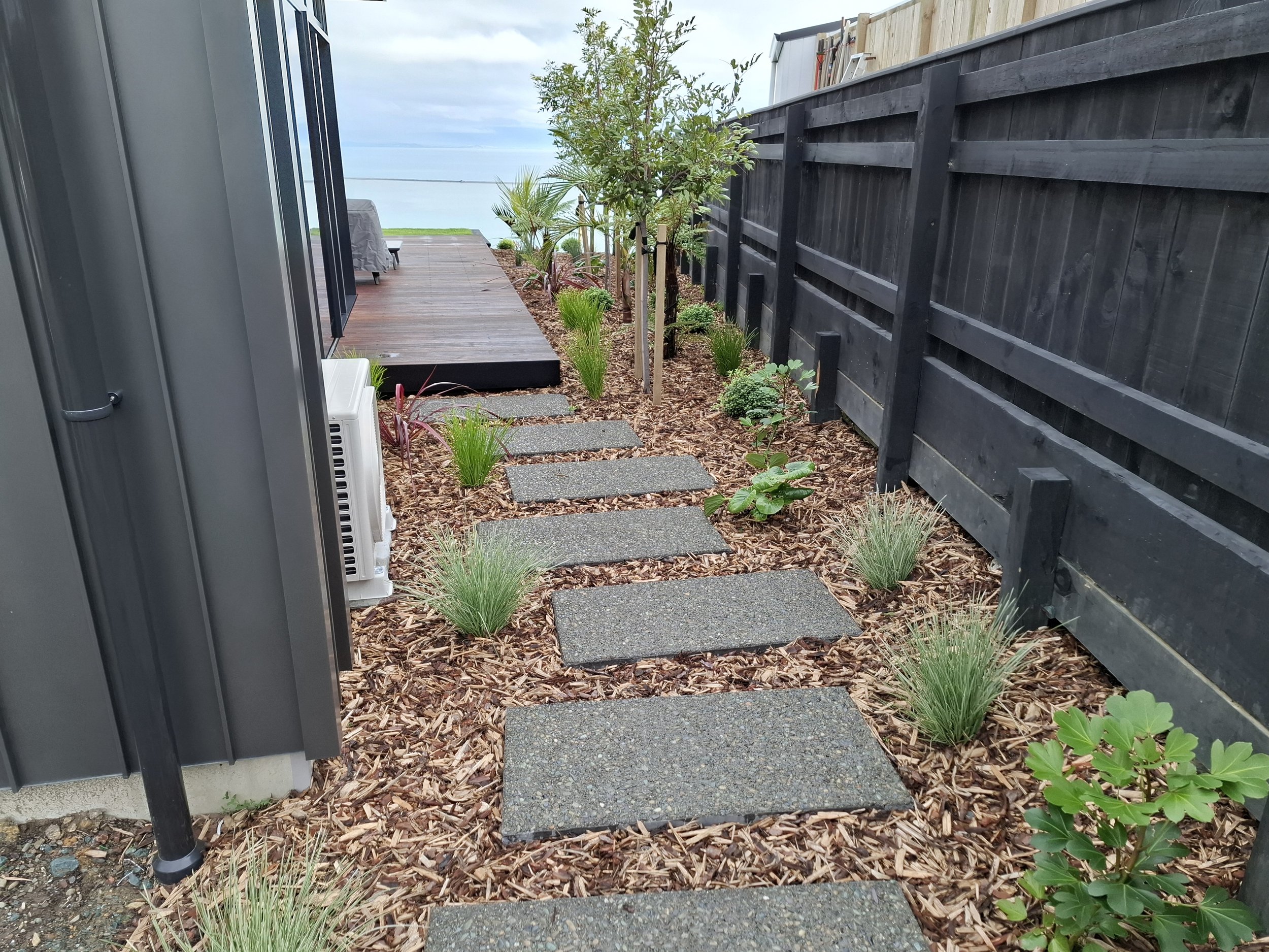 Backyard garden with stepping stones, mulch, various plants, and a wooden deck by the water, bordered by a black fence.