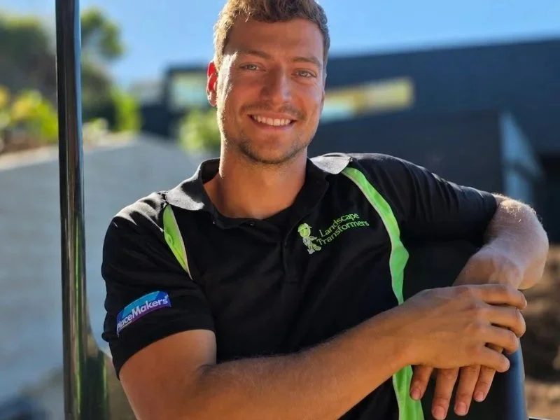 A smiling man with short, light brown hair sitting in a golf cart with a black roof, outdoors on a sunny day. He is wearing a black polo shirt with green accents and logos, including one that says 'Landscapes Transforms,' and appears to be in a landscaped area with modern buildings and greenery in the background.