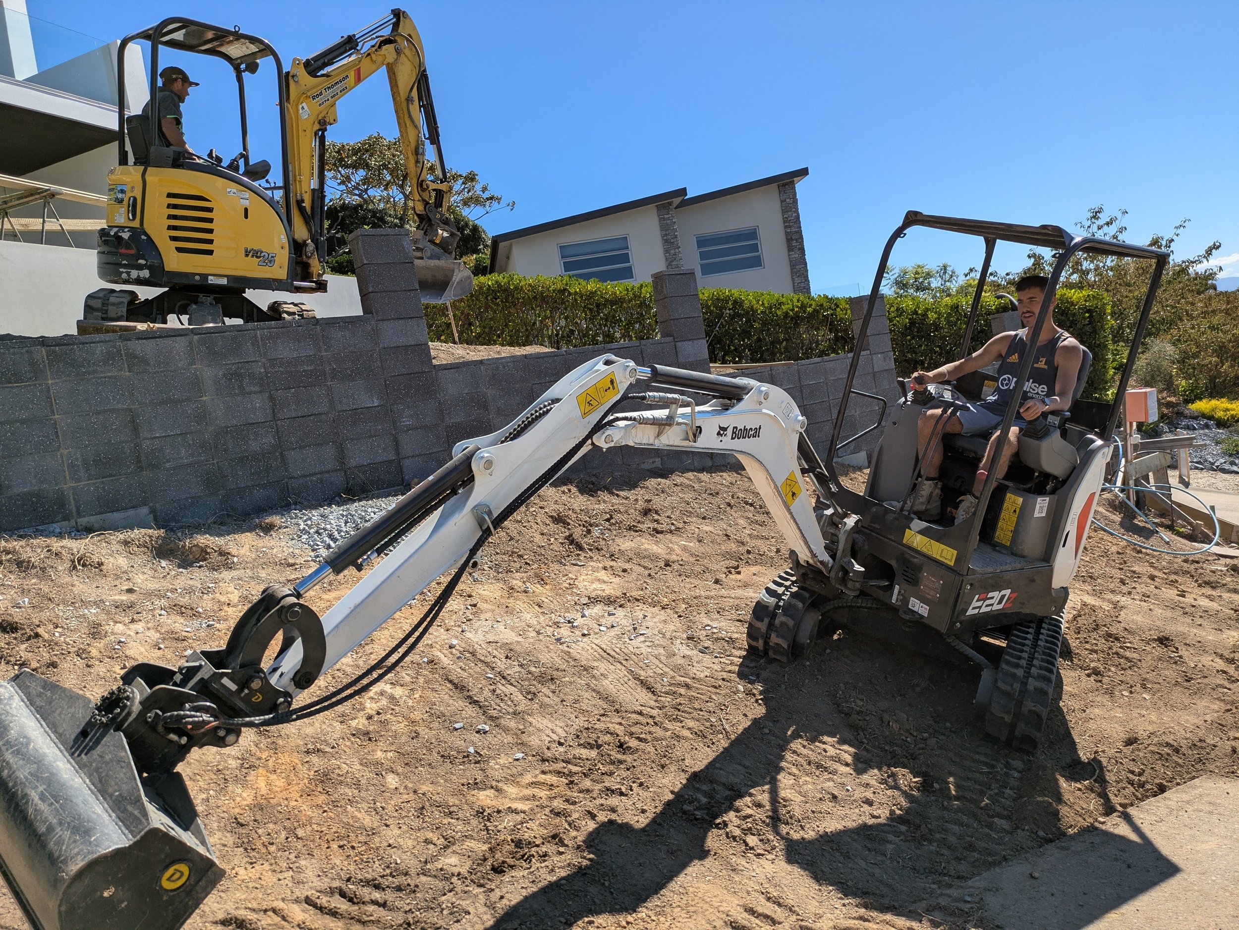 Two men operating construction equipment outdoors on a mound of dirt in a residential area with modern houses and bushes in the background.