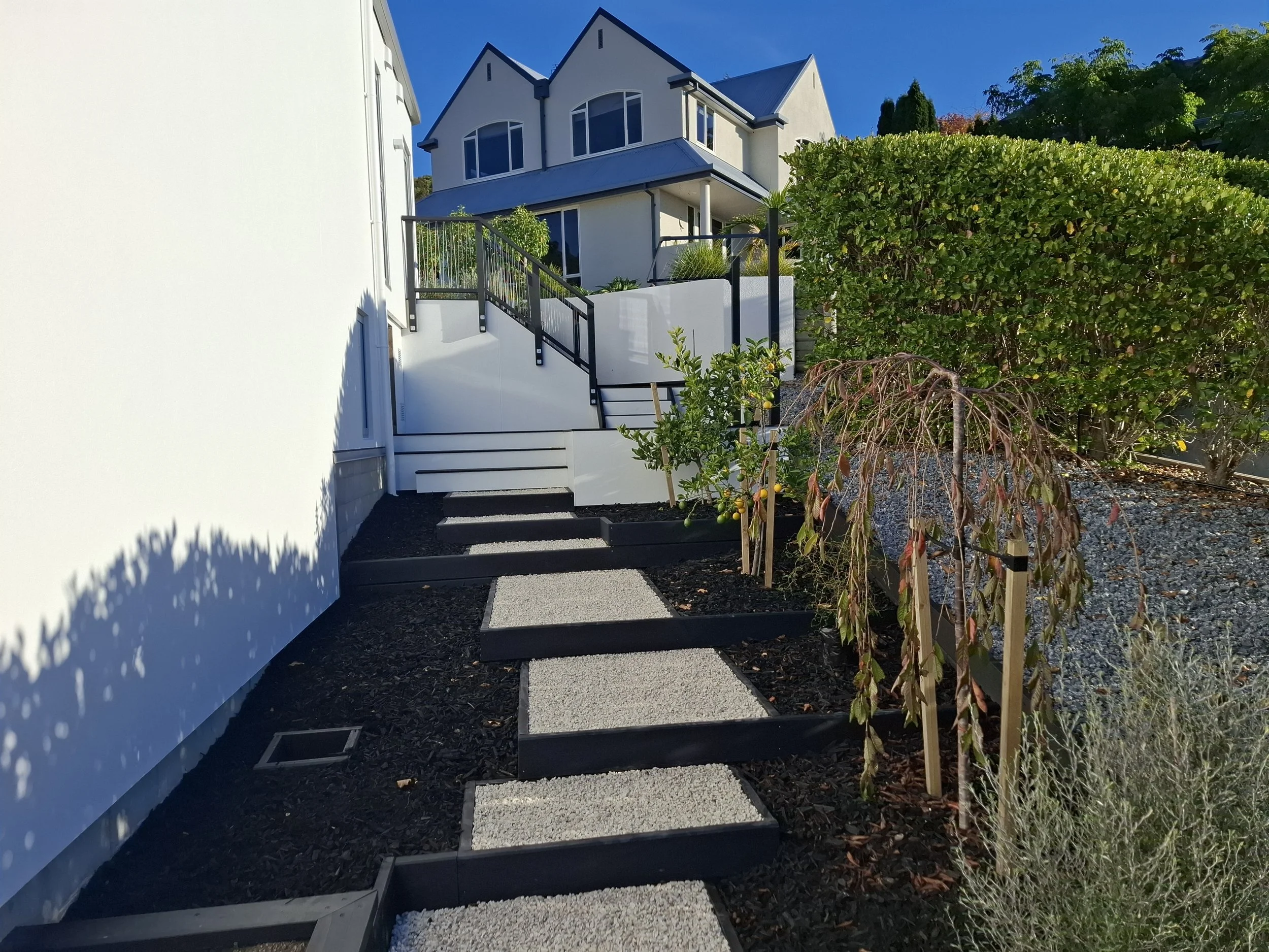 A modern house with a white exterior, black trim, and large windows, surrounded by a landscaped yard with trees and shrubs, featuring a pathway with black gravel and white stepping stones.