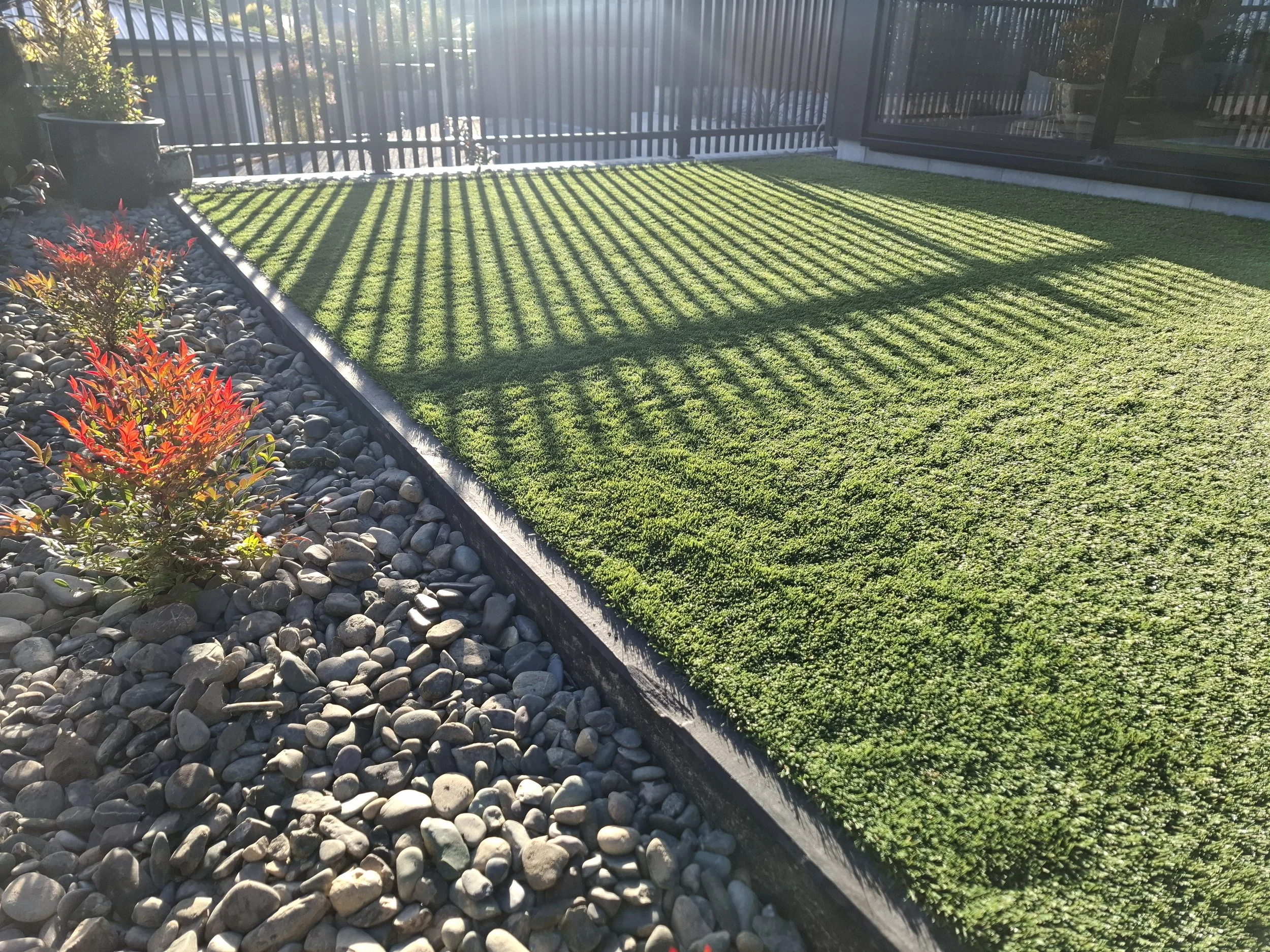 A small yard or patio area with a strip of artificial green grass separated from decorative rocks and gravel on the left side. The area is enclosed by a black metal fence and has a glass sliding door on the right side. The sunlight casts shadows from