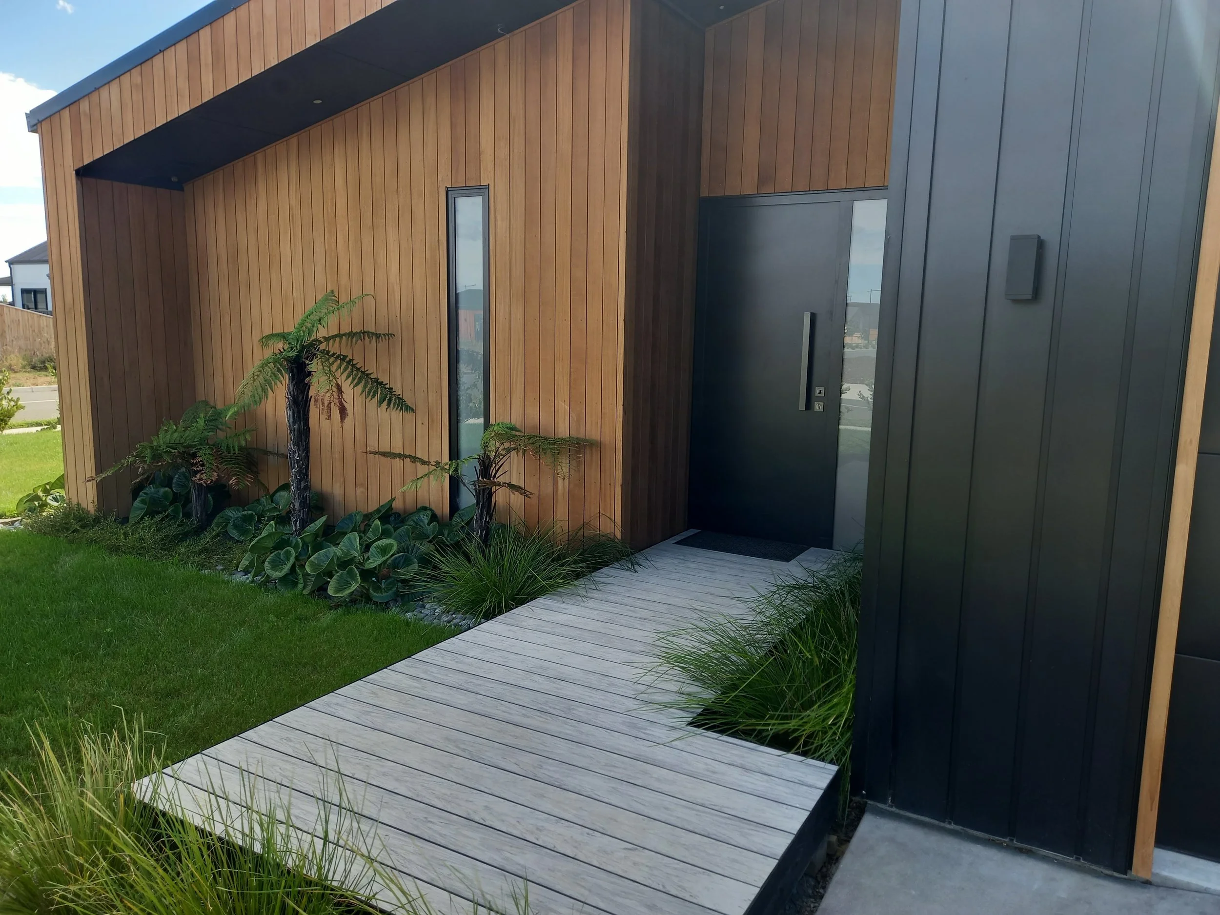 Modern house entrance with wooden and dark gray panel walls, a narrow window, a small garden with green plants, a wooden walkway, and a lawn.