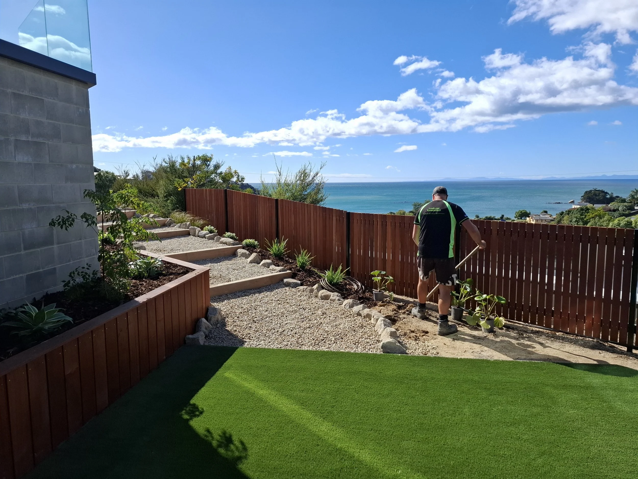 A backyard with a view of the ocean, a man working in the garden, a wooden fence, and a clear blue sky with clouds.