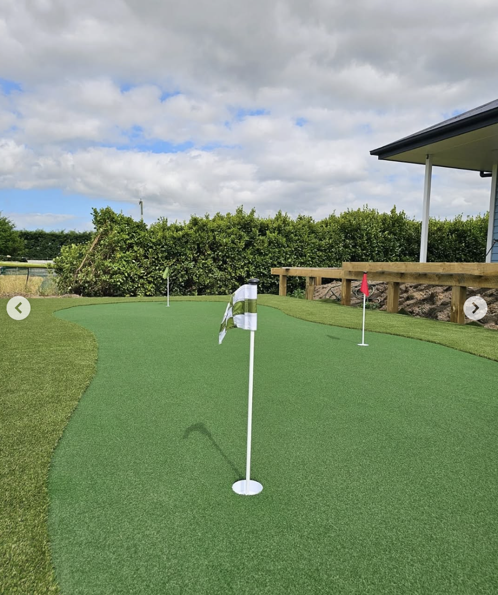 A mini golf course putting green with three flags on the holes, surrounded by bushes and a house with a porch on the right.