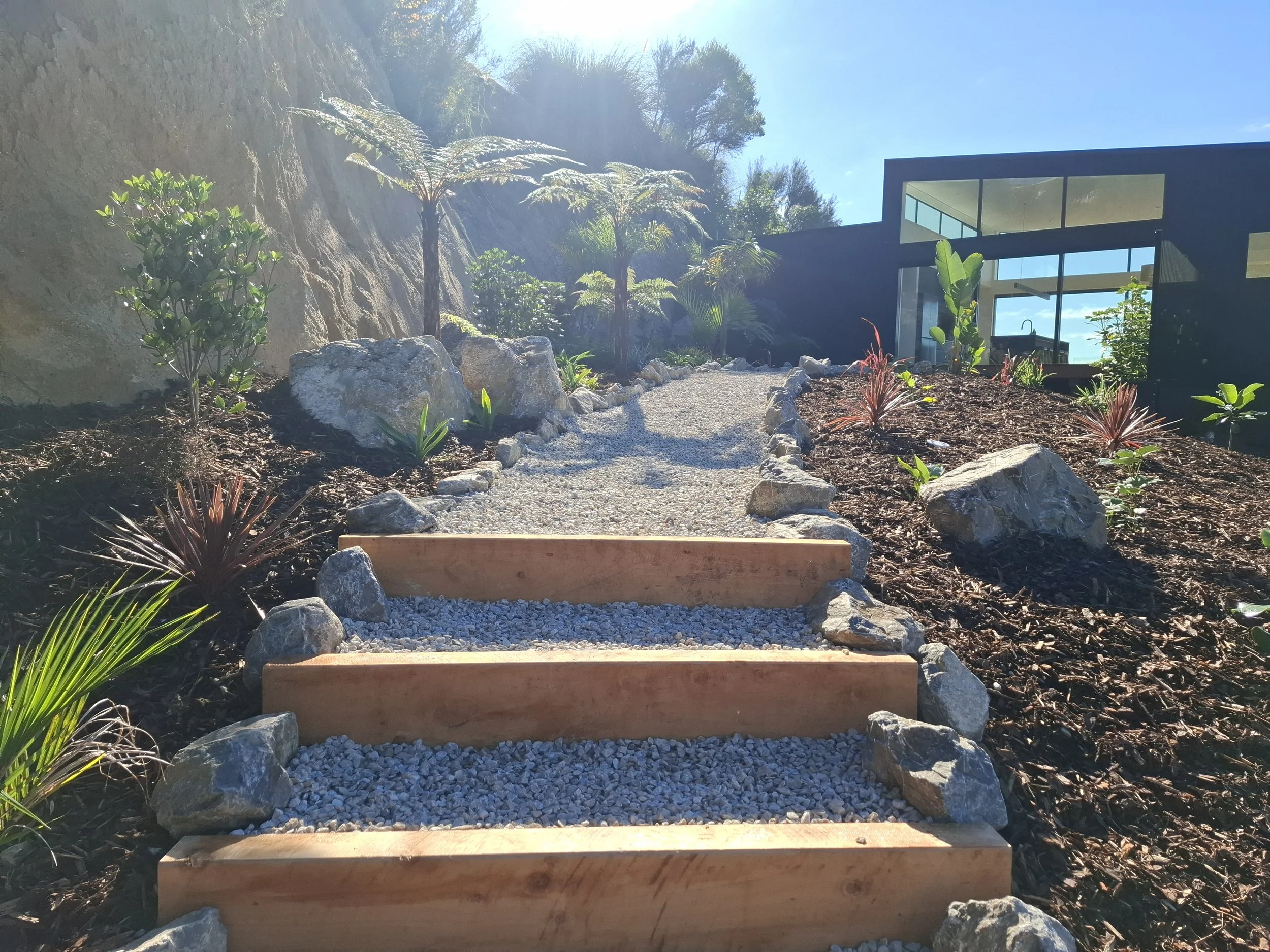 A modern house with large glass windows is set against a hillside with lush plants and trees. There are stone steps and a gravel pathway leading up to the house, with greenery and rocks surrounding the area. The sky is clear and blue, indicating a su