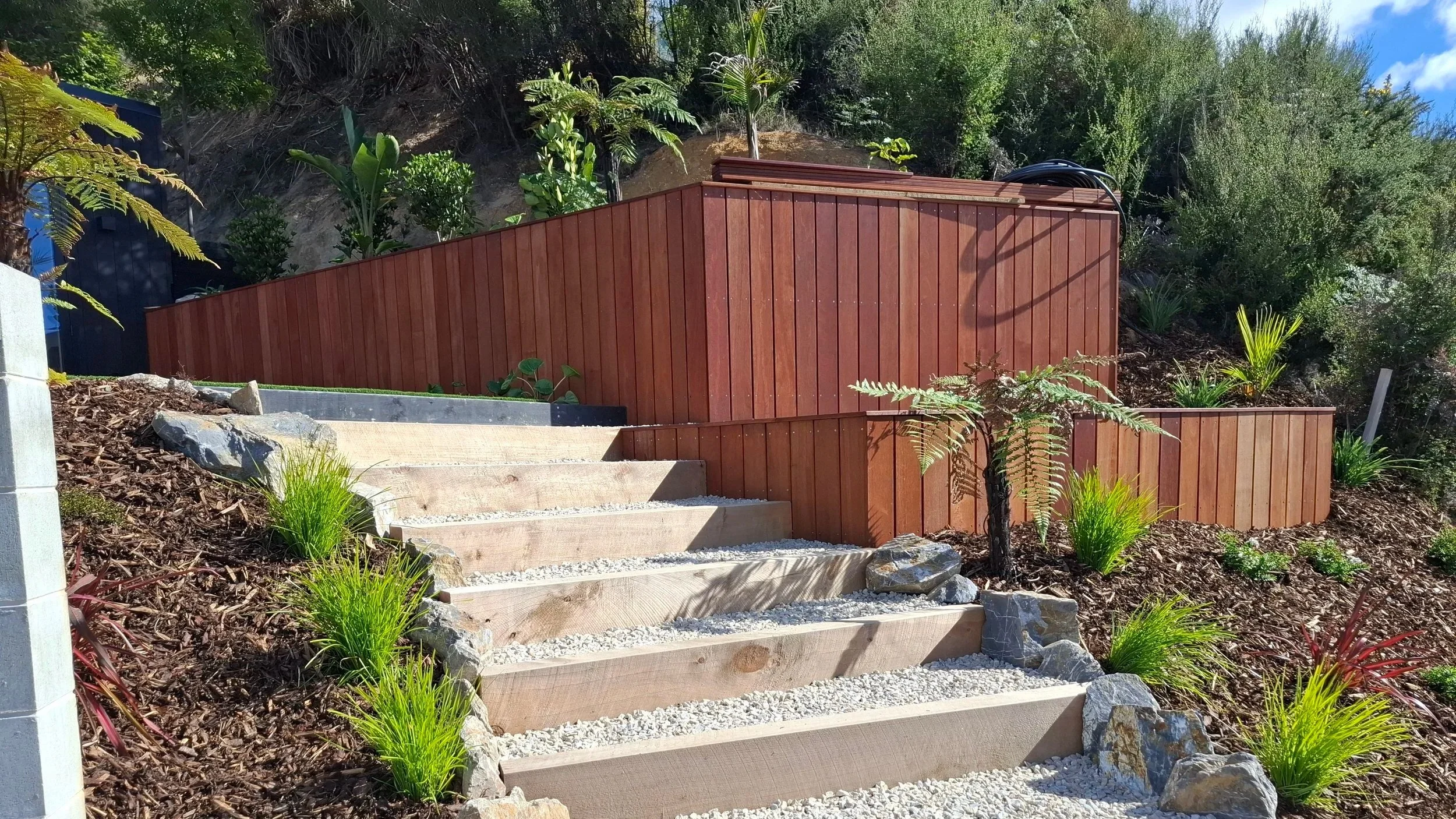 Outdoor garden area with stone stairs, a dark red wooden fence, and lush green plants on a slope.