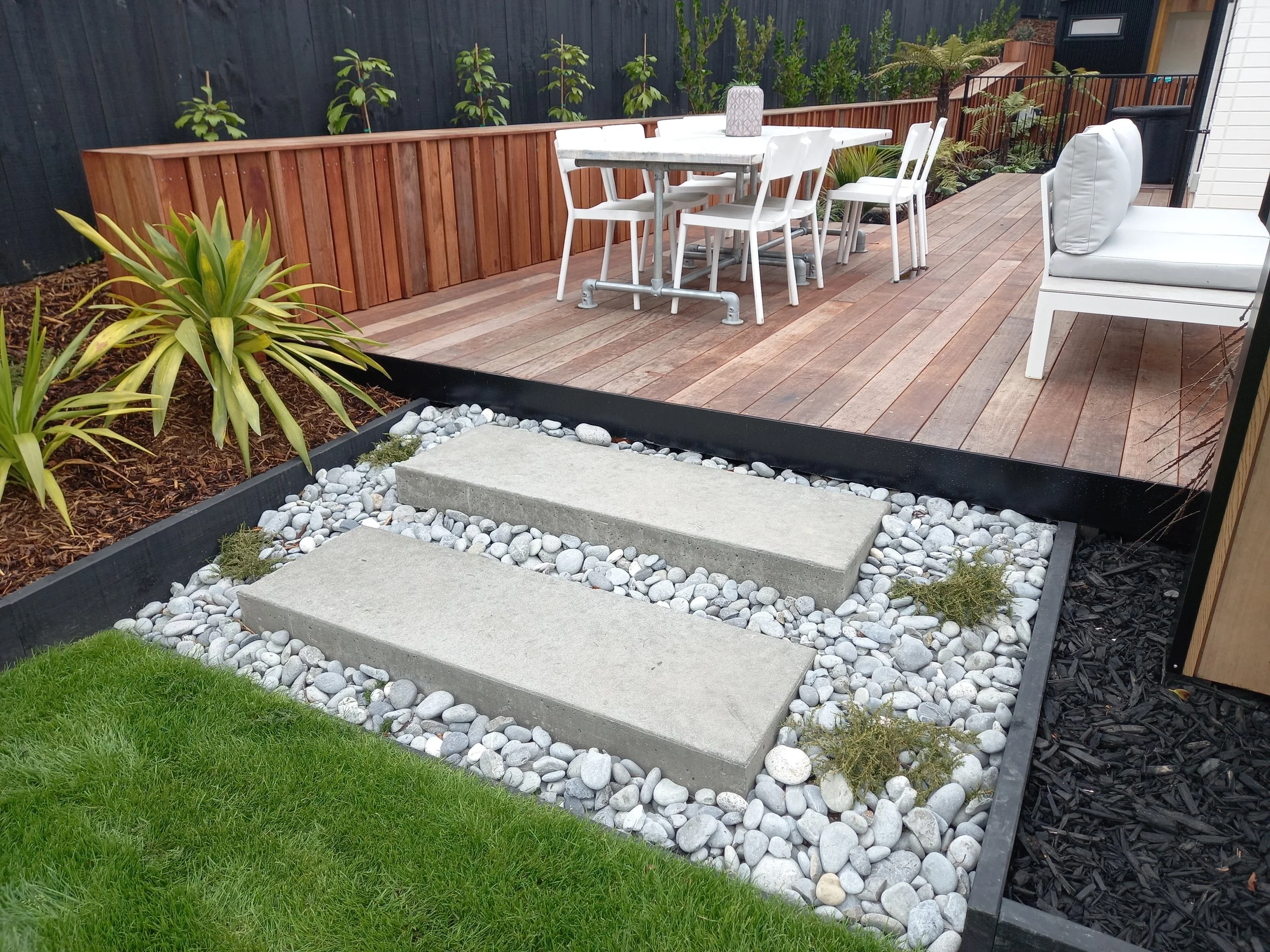 A backyard patio with a wooden deck, white outdoor furniture including a table and chairs, surrounded by plants, a black privacy fence, and decorative rocks and steps leading up to the deck.