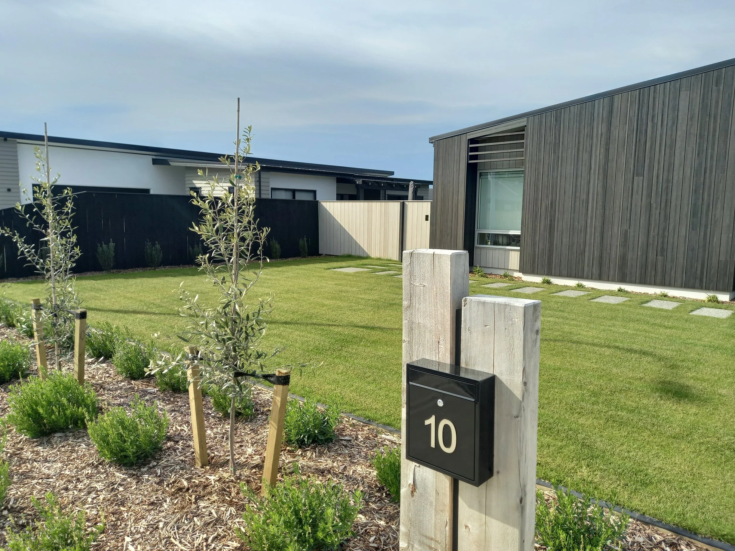A modern residential backyard with a neatly maintained lawn, small trees, and bushes, surrounded by a black privacy fence. In the background, there are contemporary houses with clean lines and large windows. A wooden mailbox with the house number 10 