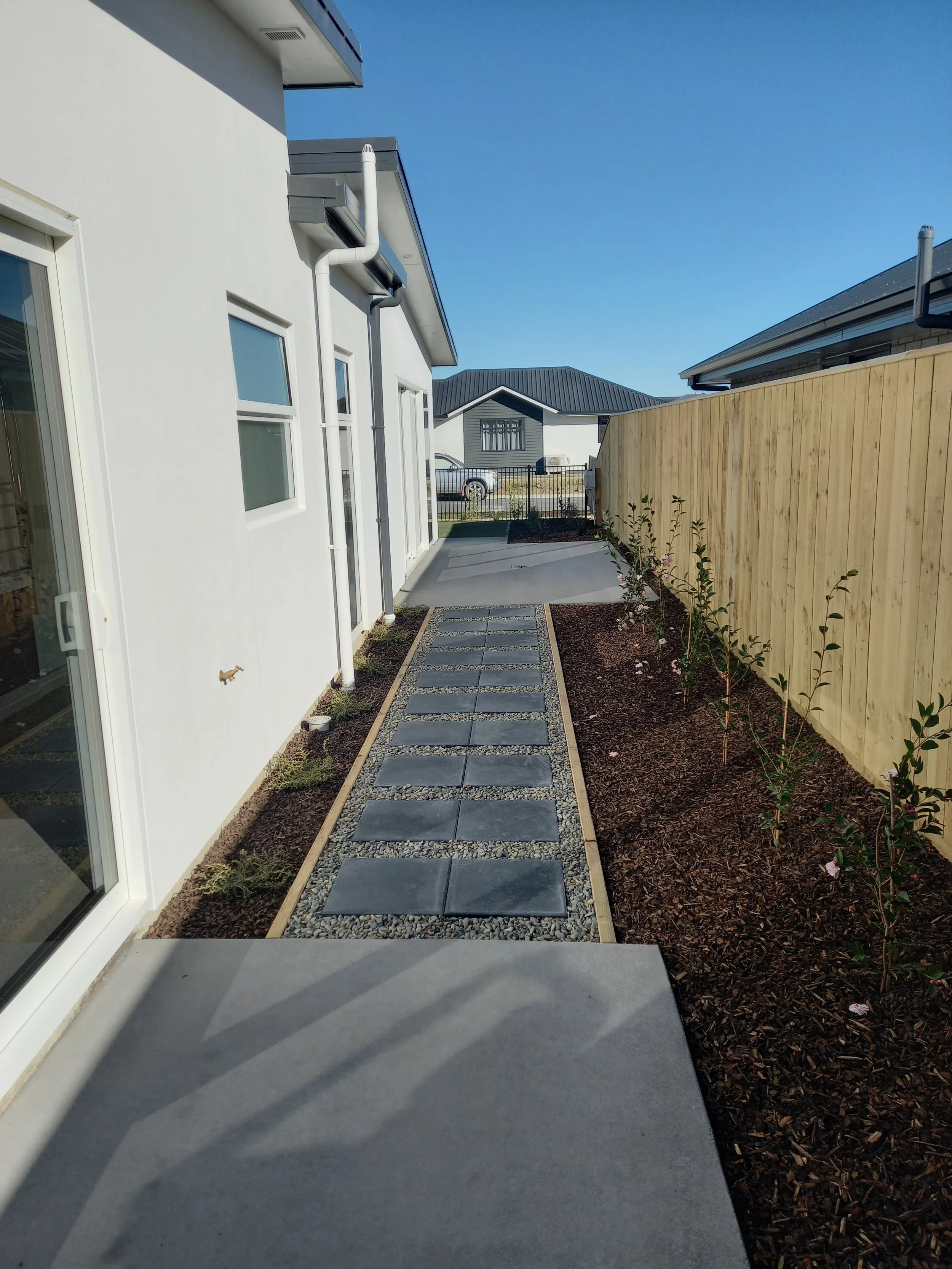 A narrow backyard pathway with large gray stepping stones bordered by gravel, bordered on the right by a wooden privacy fence with small plants growing alongside, and on the left by the exterior wall of a white house with large glass sliding door and