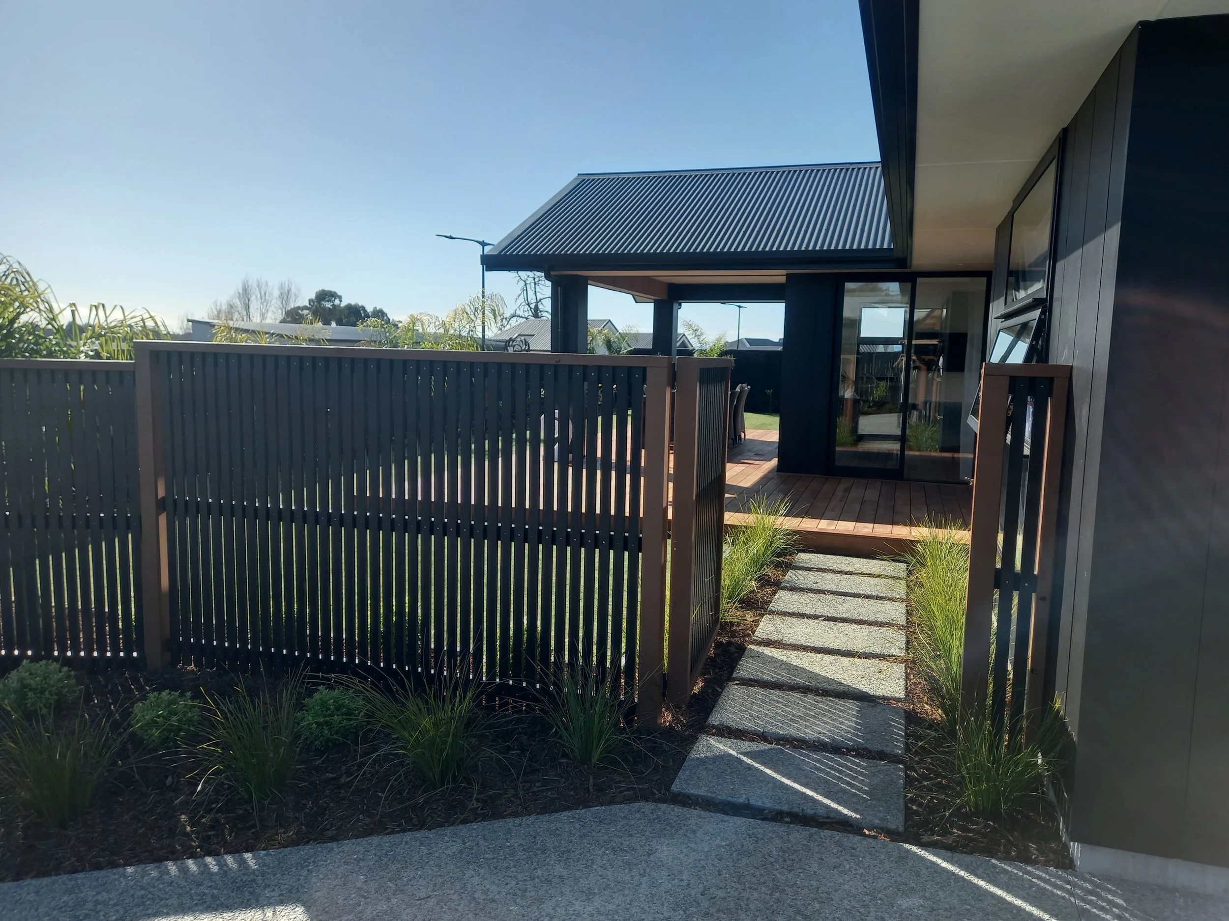 Modern house with dark exterior, large glass sliding door, and a small patio with a wooden fence and stepping stones, surrounded by grass and plants.