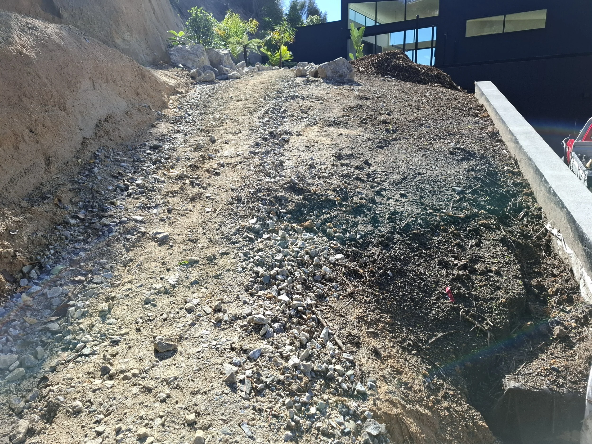 A dirt and gravel pathway beside a modern black building with large windows, with plants and rocks at the edge.