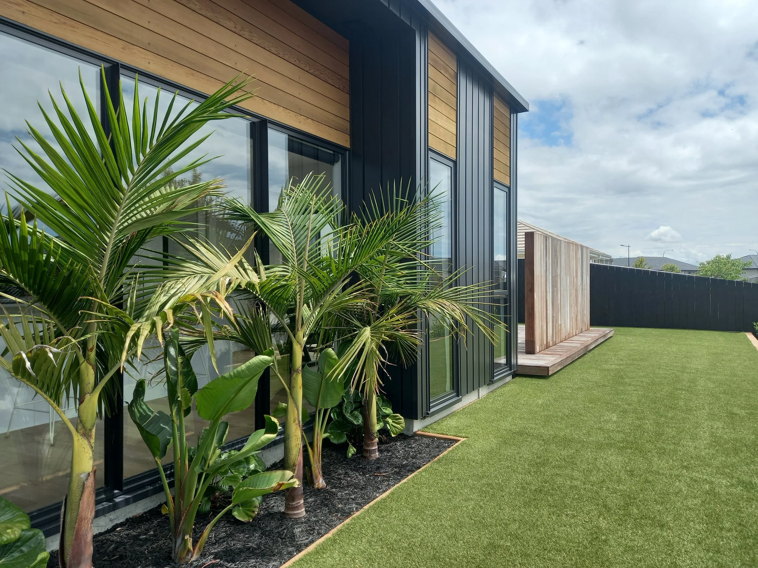Modern house with black metal siding and wooden accents, large glass windows, and a well-maintained lawn with tropical plants.