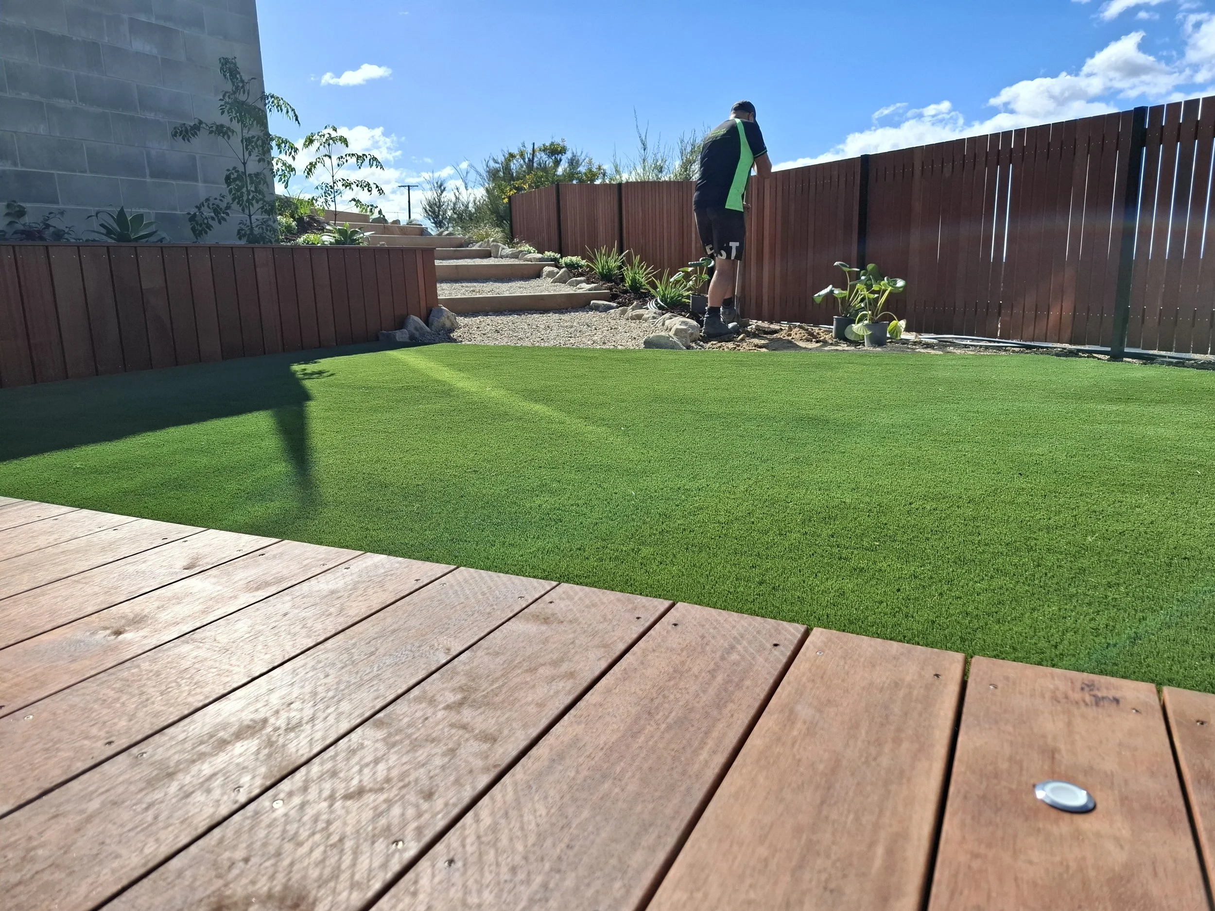A man is working on a garden with a wooden fence, plants, and a green artificial grass yard on a sunny day with a blue sky and clouds.