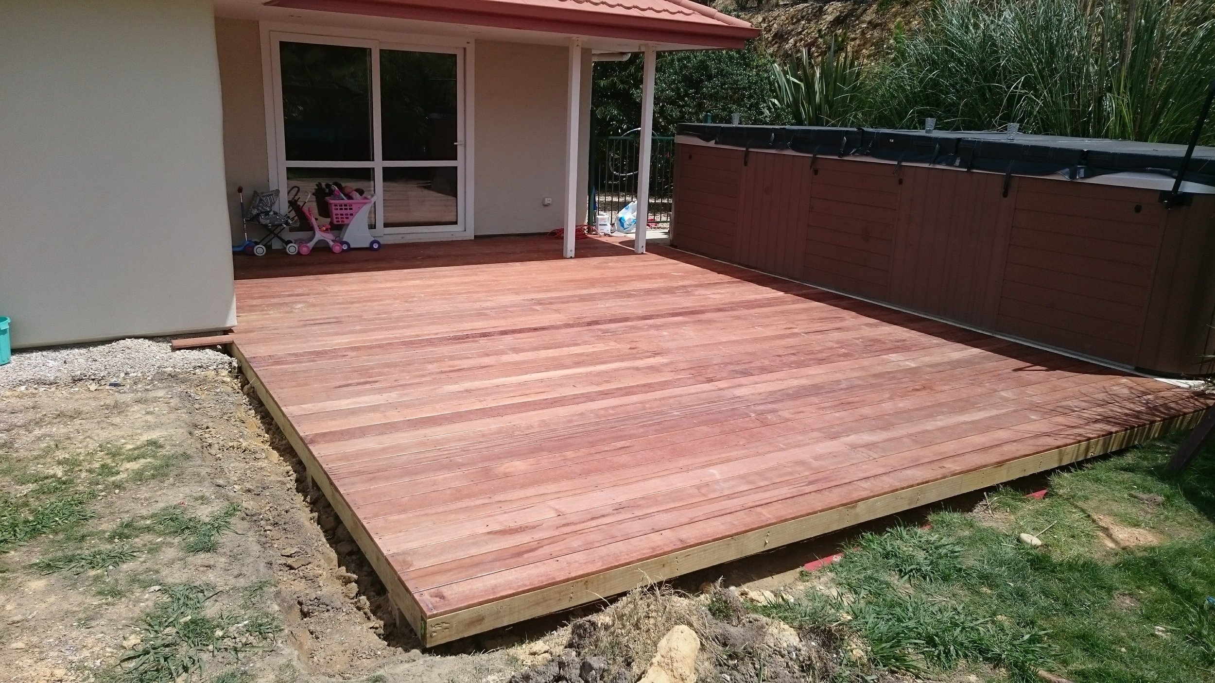 Newly installed wooden deck outside a house with sliding glass door, next to a hot tub, surrounded by grass and garden.