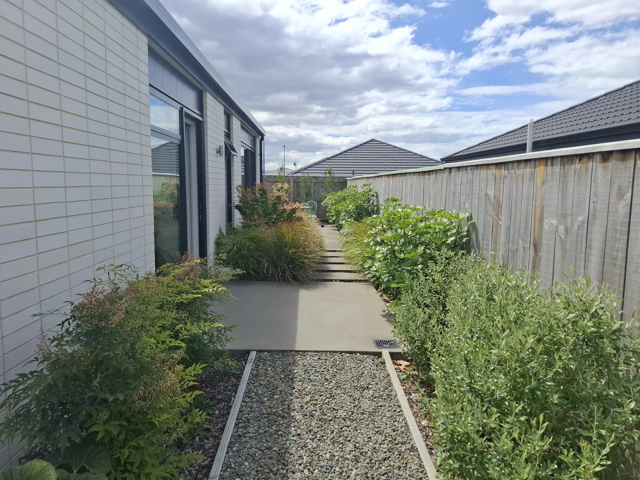 A small garden path lined with green shrubs and plants, with a wooden fence on the right and a modern house with large windows on the left, under a partly cloudy sky.