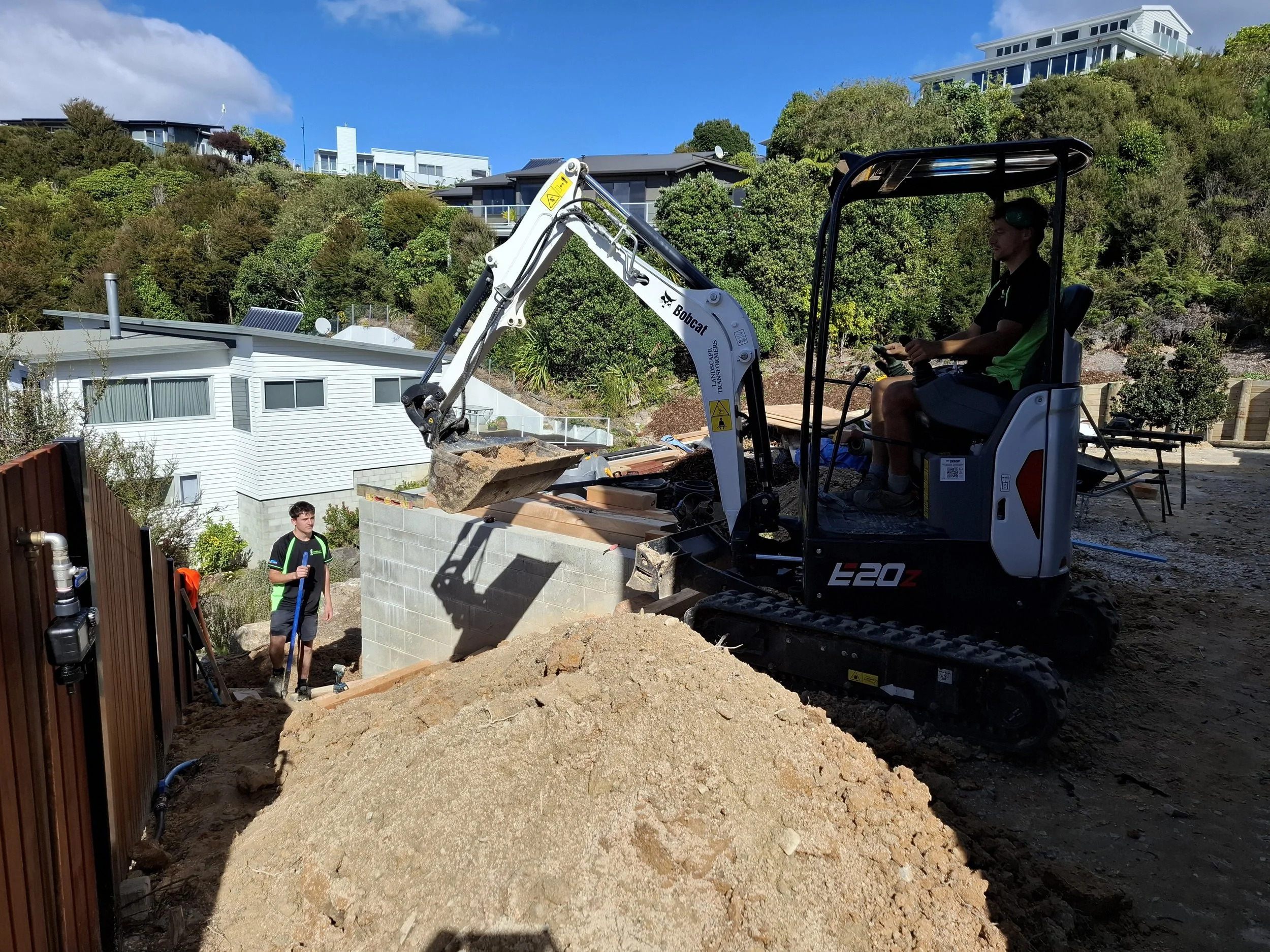 A man operating a mini excavator on a construction site in a residential area with houses and greenery in the background.