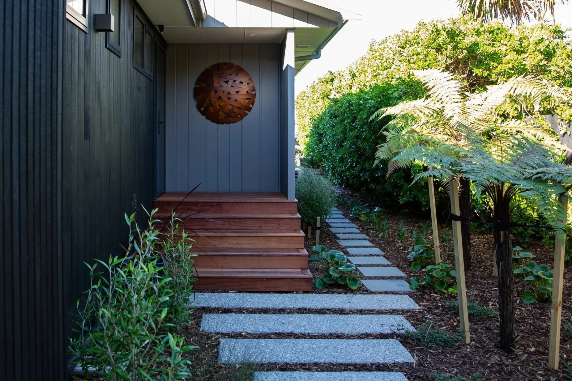 A pathway made of square stone pavers leading to wooden stairs at a modern house entrance. The house has a dark exterior wall with a circular wooden art piece. Lush green plants and trees line both sides of the pathway, creating a garden setting.