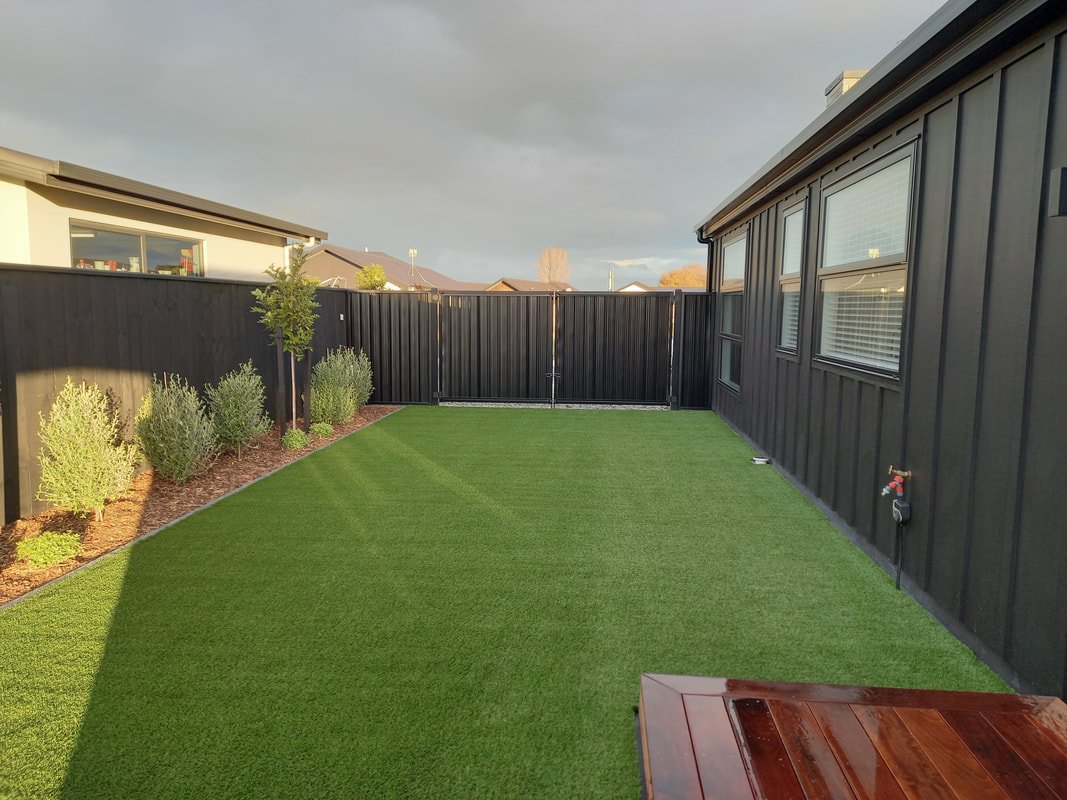 A fenced backyard with a neatly mowed green lawn, small trees and shrubs along the left side, black metal fencing, and windows on a black building on the right side, with a wooden table in the foreground.