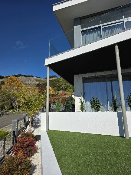 Modern house with large glass windows, balcony, and a well-maintained lawn, situated in a hilly area with colorful trees and blue sky in the background.