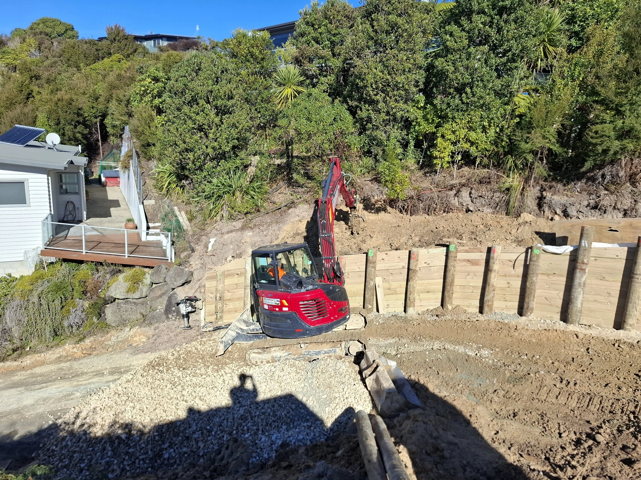 Construction site with a small red excavator working on a dirt and gravel area, a wooden fence, and lush green trees in the background.