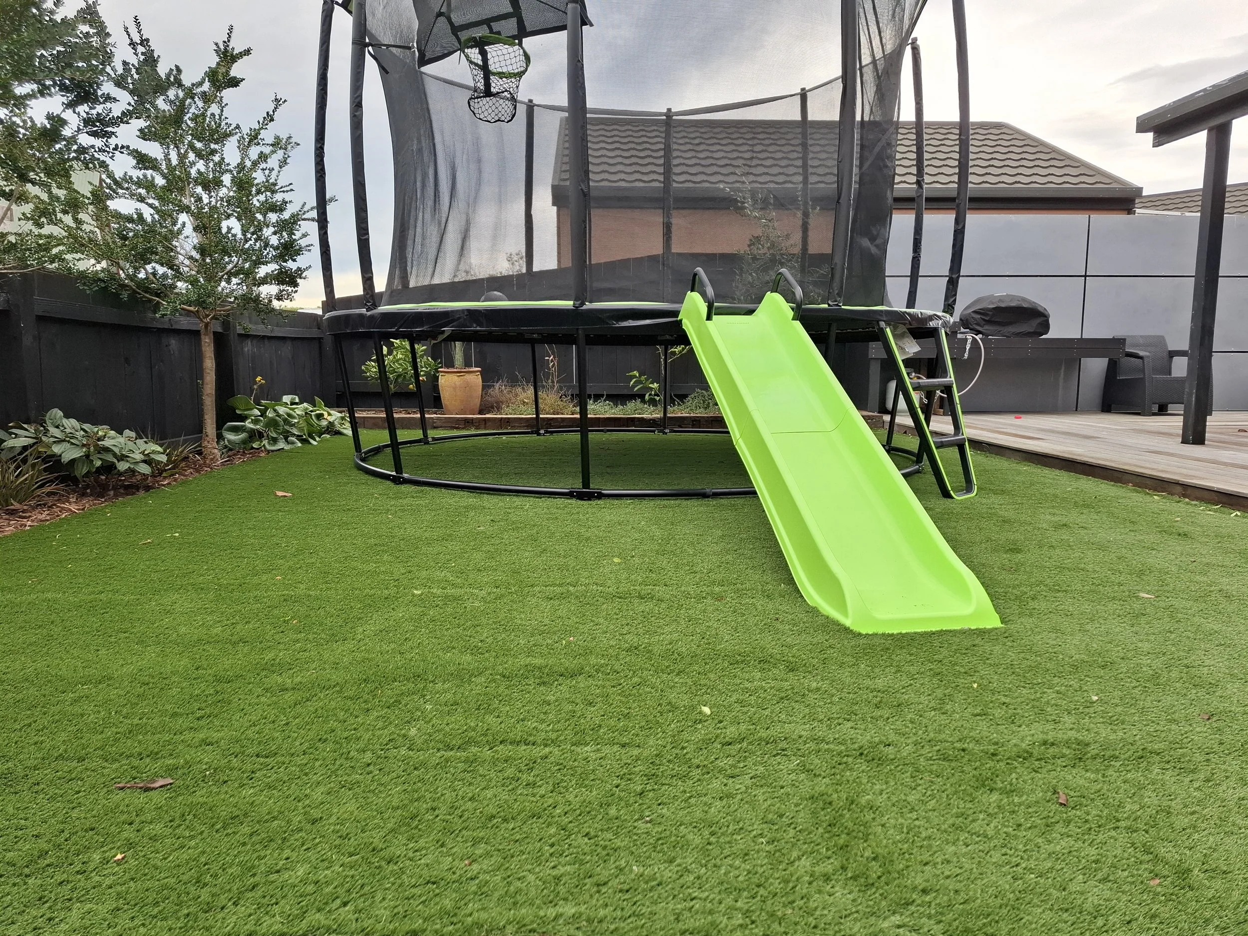 Backyard with artificial grass, a trampoline with a green slide, some plants, and a tree, with a house and fence in the background.