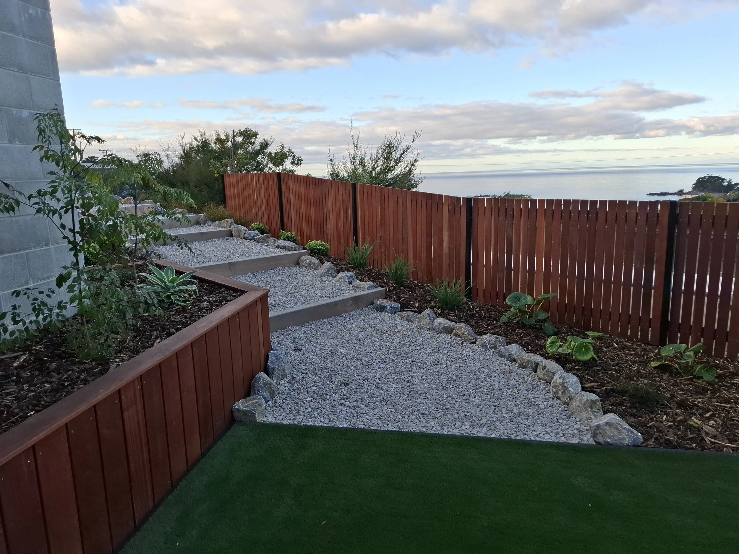 A backyard garden with a view of the ocean and cloudy sky. There is a red wooden fence, landscaped plants, and rocky pathways.