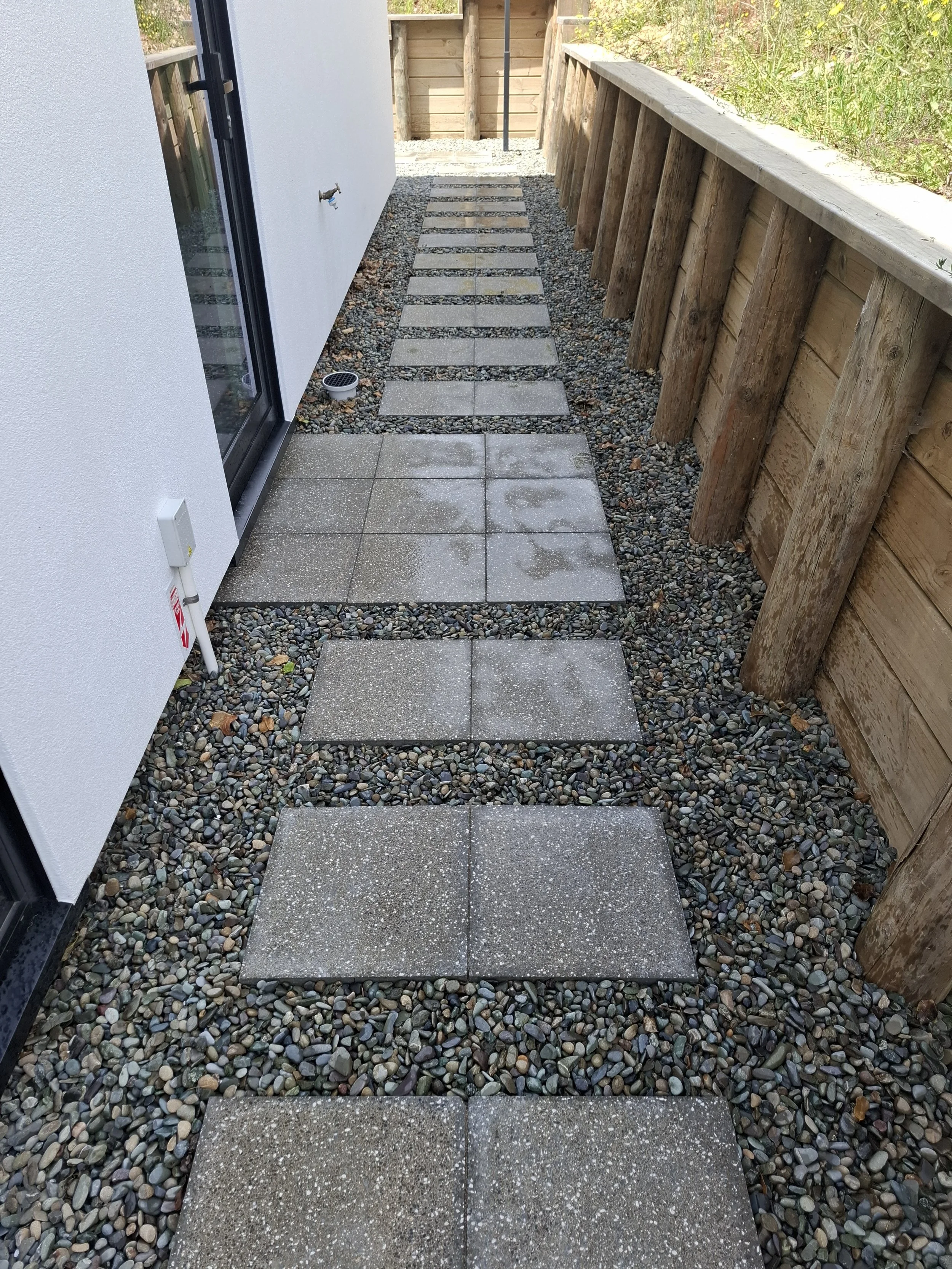A narrow outdoor pathway with rectangular concrete pavers surrounded by small multicolored pebbles, leading to a wooden fence at the back, with a wooden fence on the right side and a white wall of a building on the left.