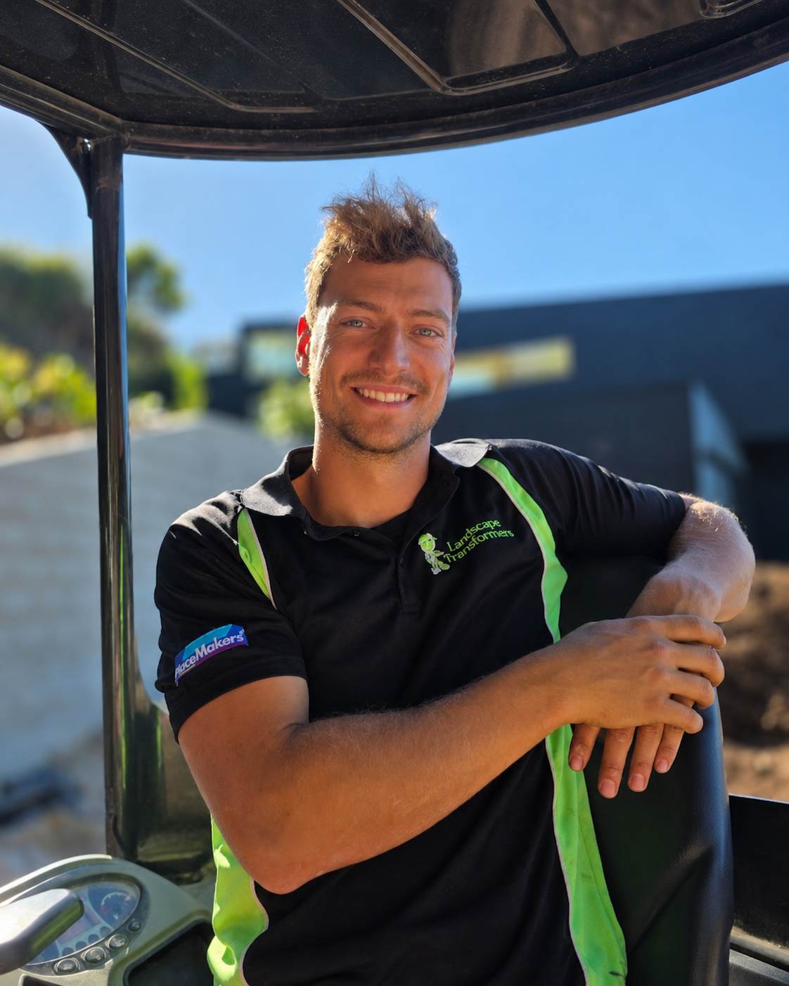 A smiling man sitting in a golf cart outdoors with a modern building and trees in the background, wearing a black and green polo shirt with logos.