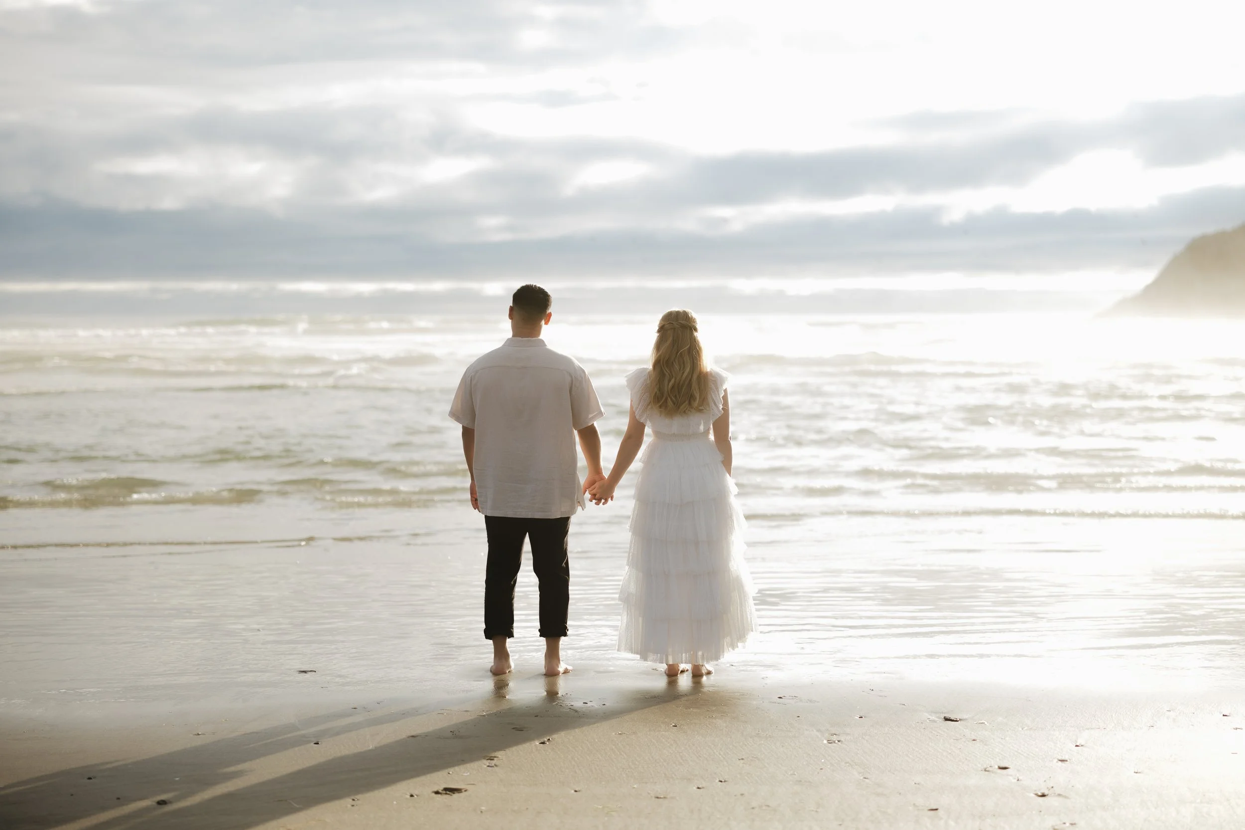 A couple holding hands, standing barefoot on the beach facing the ocean, with cloudy skies above.