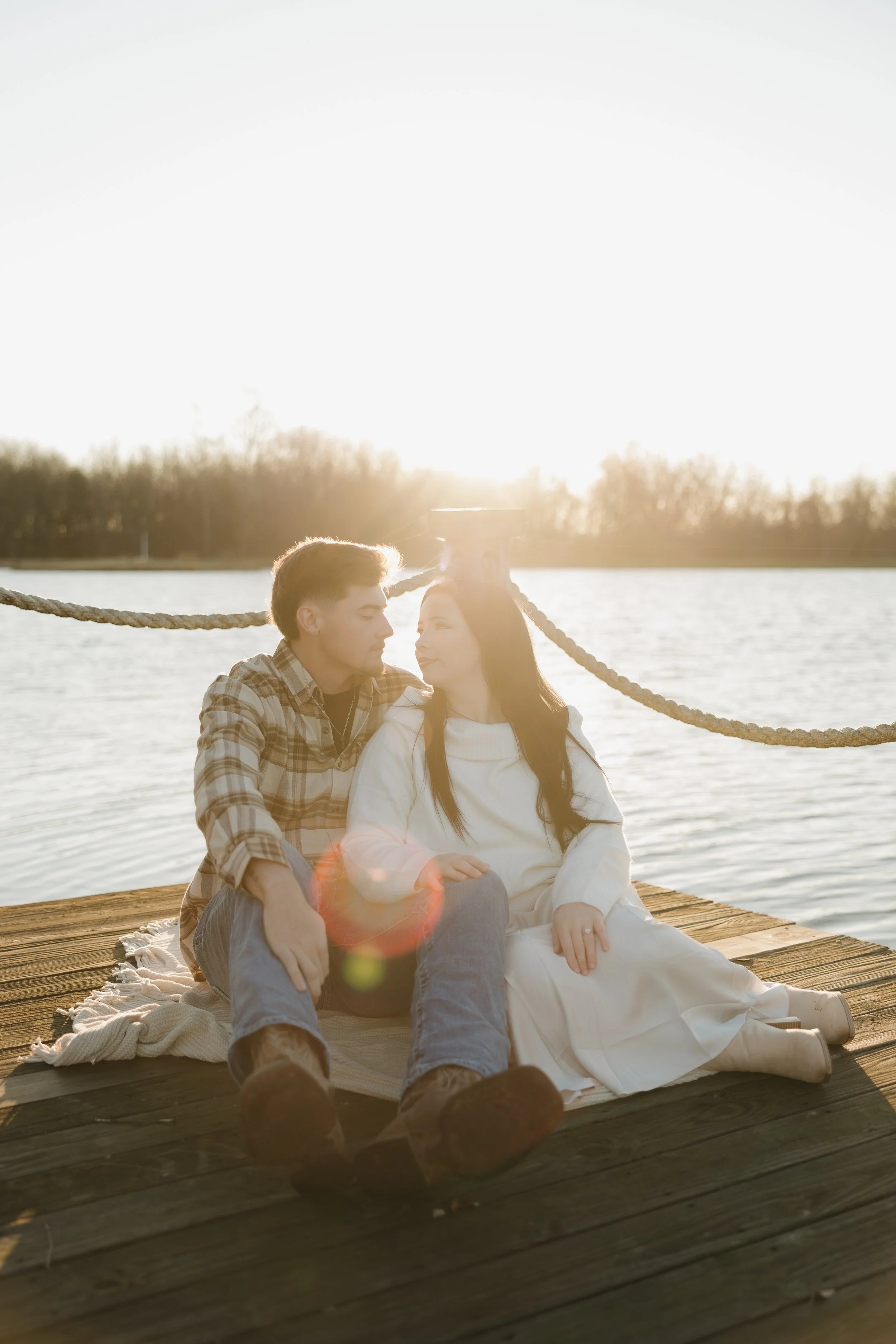 A young couple sitting on a dock by a lake during sunset, looking at each other affectionately with sunlight shining behind them.