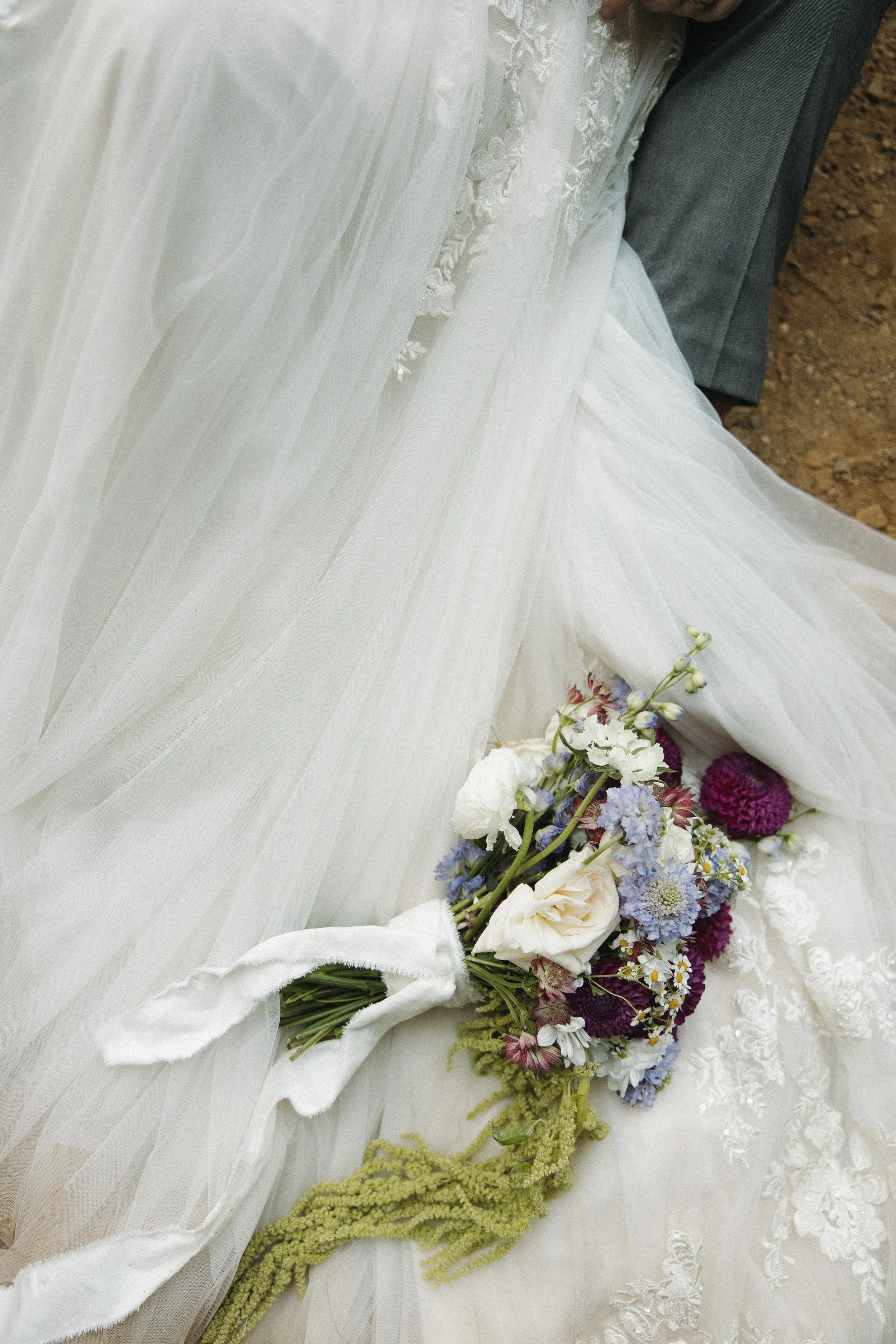 Close-up of a wedding dress with lace details, a bouquet of white, purple, and blue flowers, and a white ribbon, with part of a person's grey suit visible.