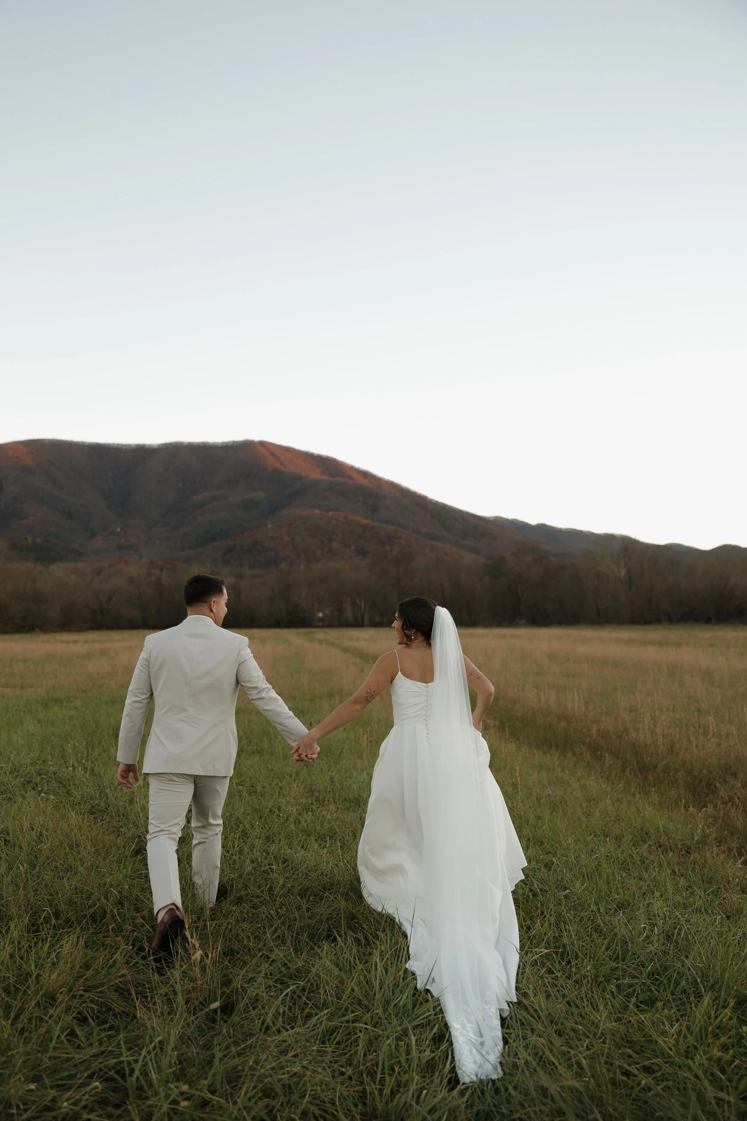 A bride and groom holding hands while walking in a grassy field at sunset, with mountains in the background.