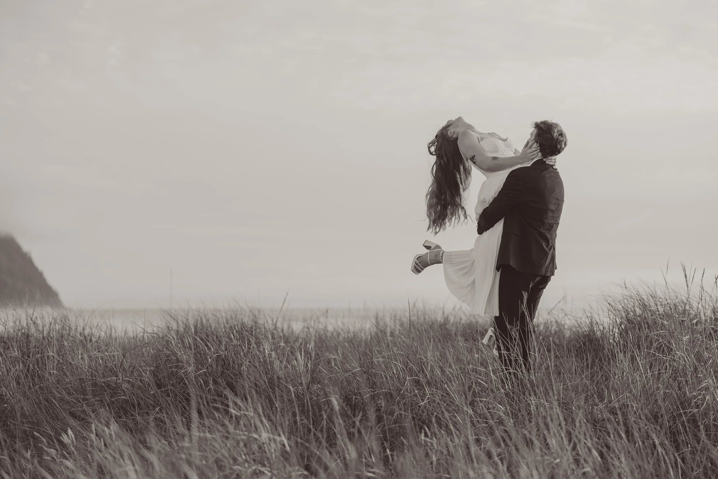 A man in a suit lifting a woman in a white dress in a grassy field, with a distant shoreline and a hill in the background.