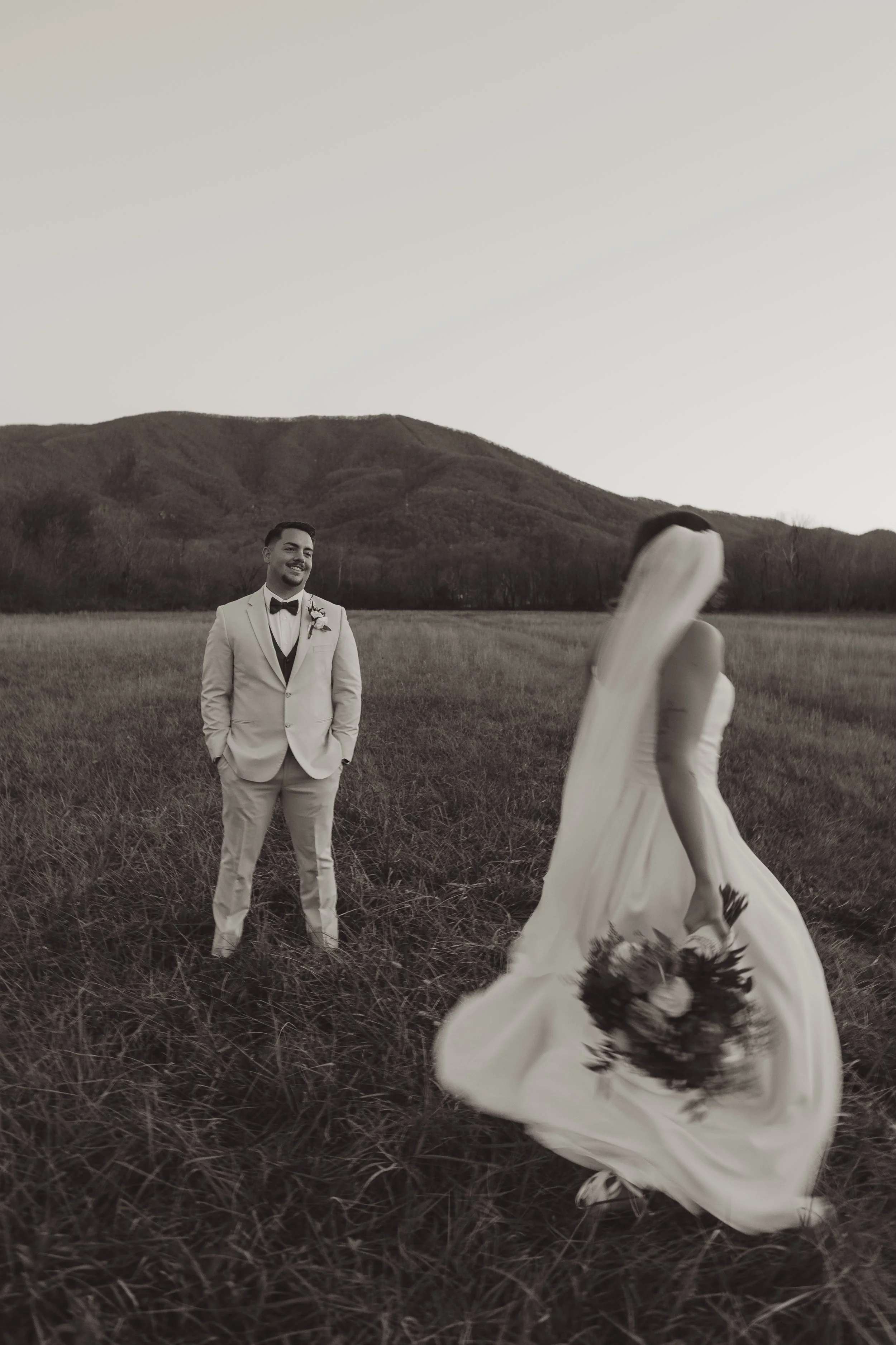 A groom in a white suit and bowtie standing in a field, smiling at a bride whose wedding dress and bouquet are caught in the wind, with mountains in the background during sunset.