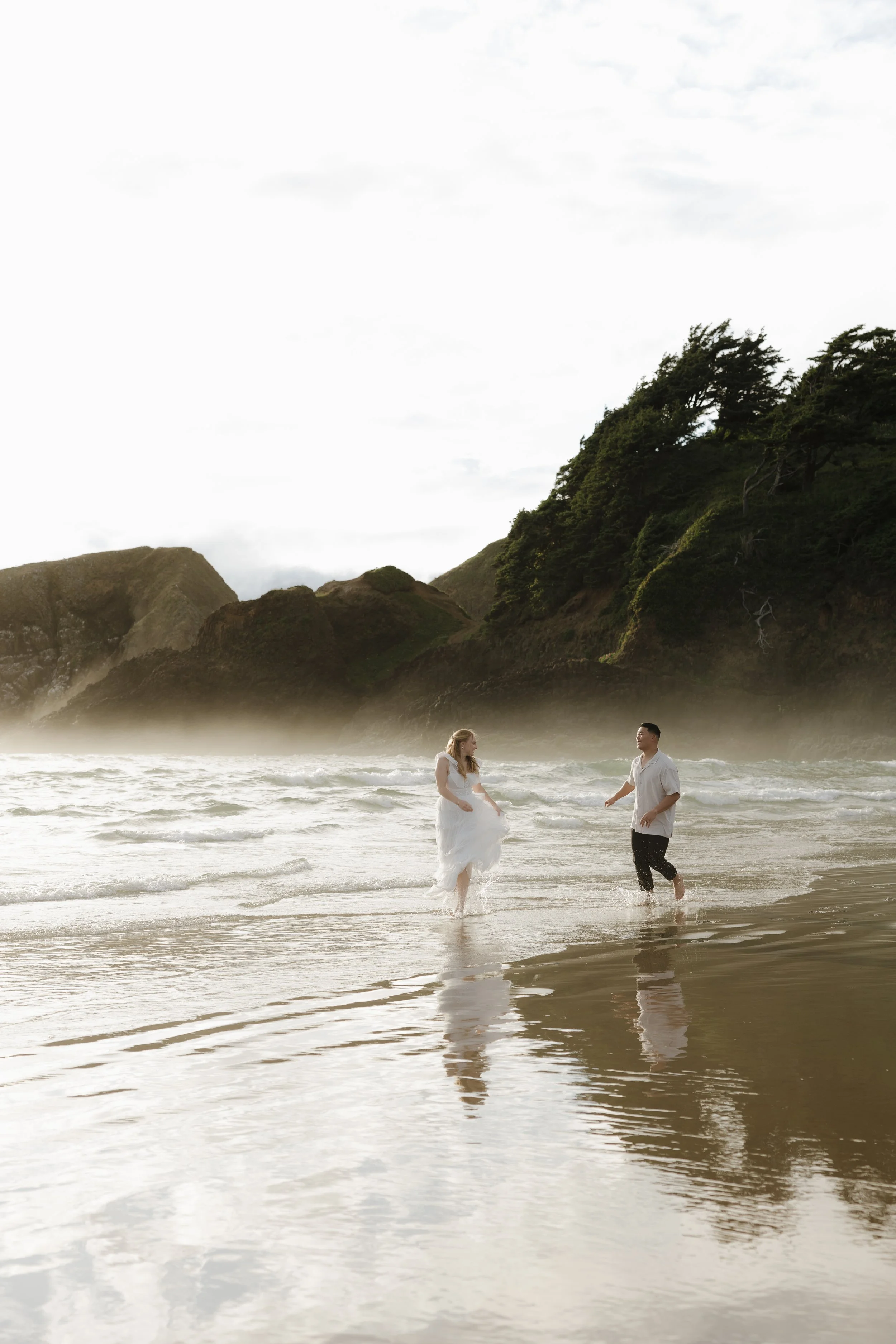 A couple in wedding attire walking in the shallow surf on a beach with green hills and mist in the background.