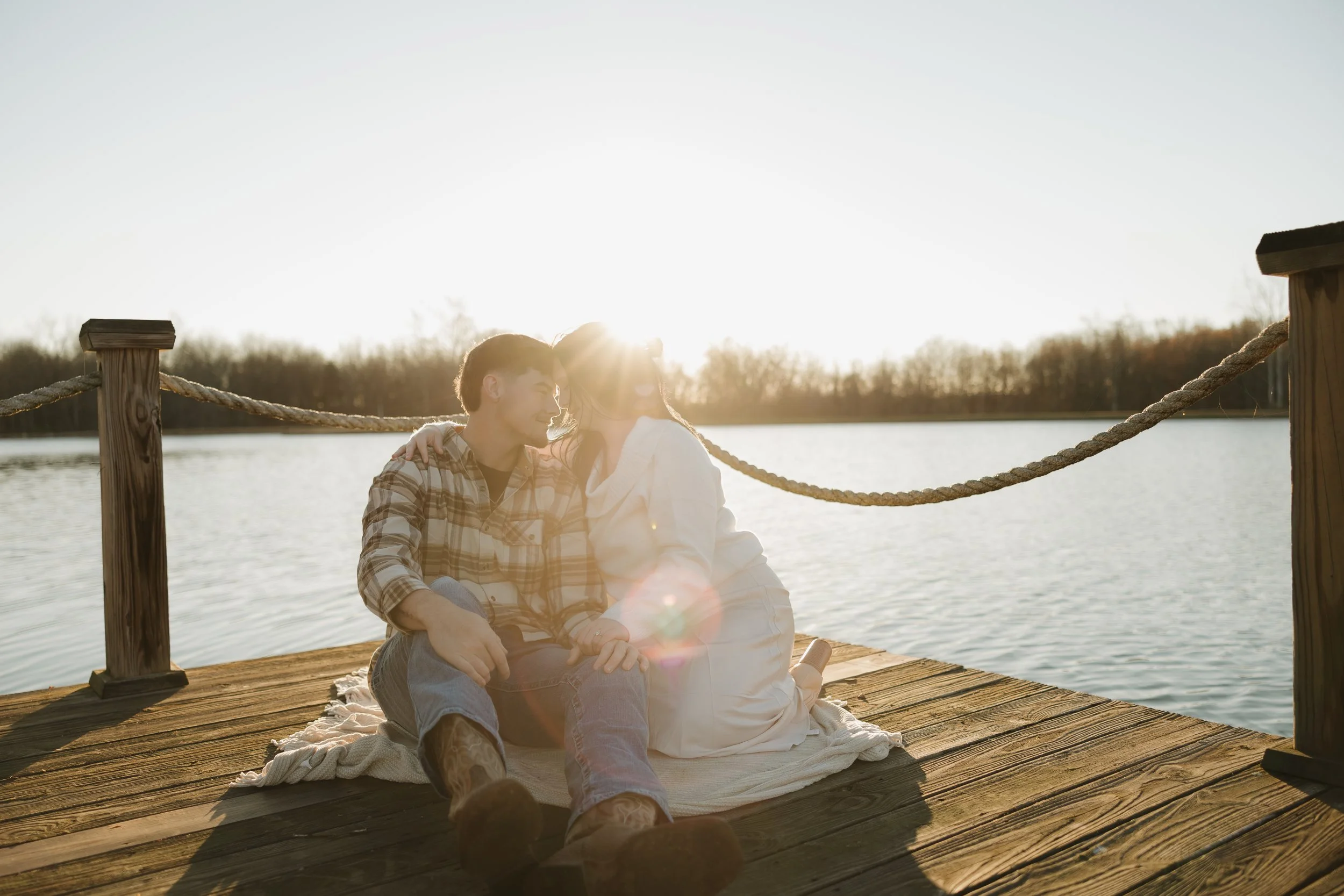 A couple sitting on a wooden dock by the water, holding hands, with the sun setting behind them.