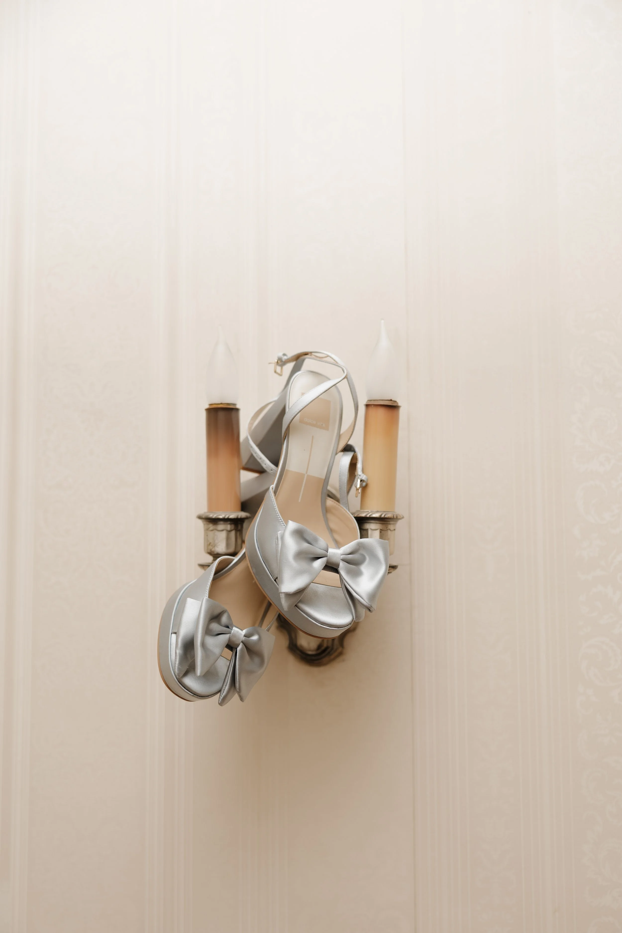 Silver high-heeled sandals with large bows hanging on a wall sconce with two candle-shaped bulbs.