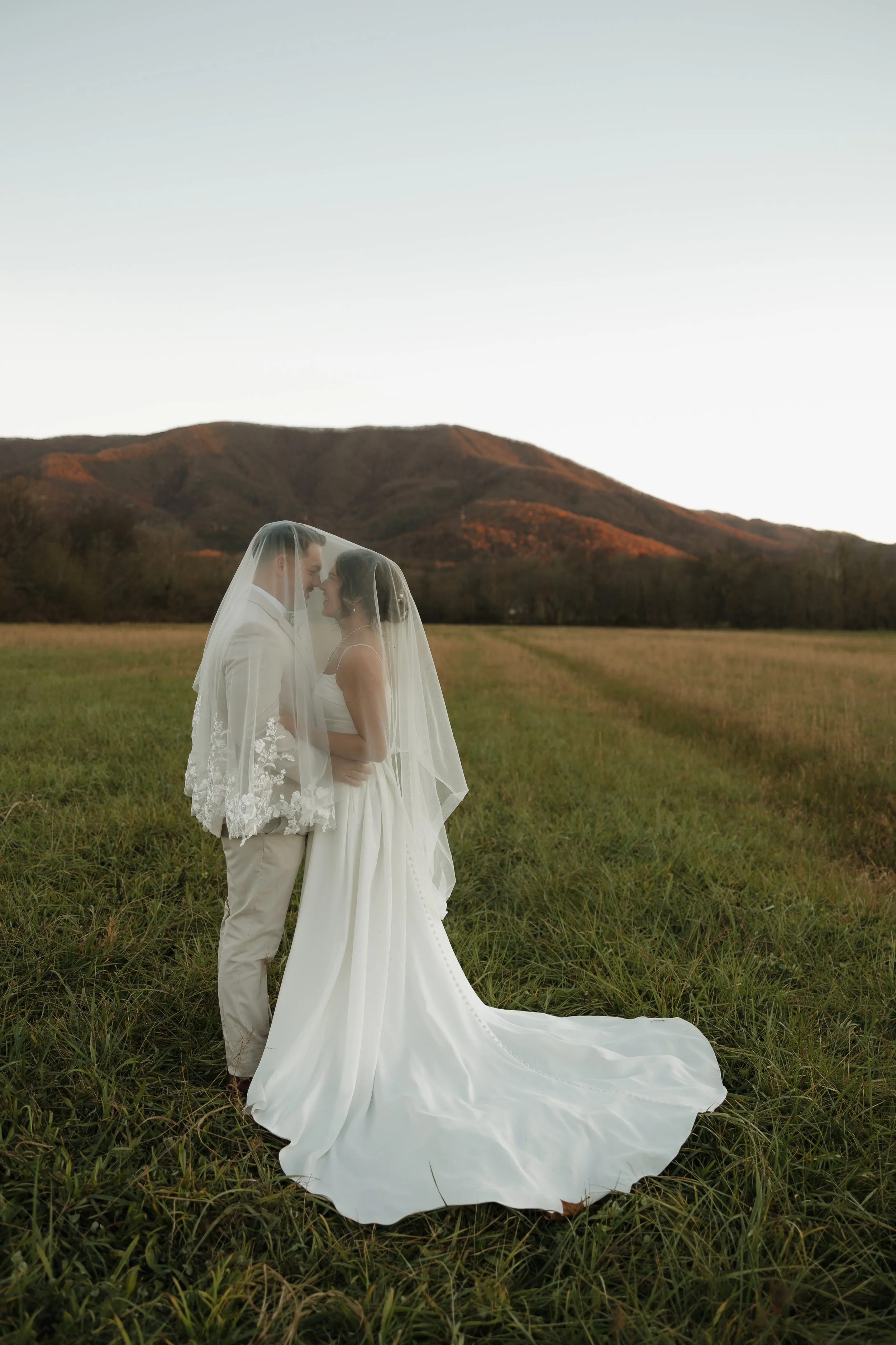 A bride and groom standing close together, sharing a moment under a veil, in an open field with mountains in the background during sunset.